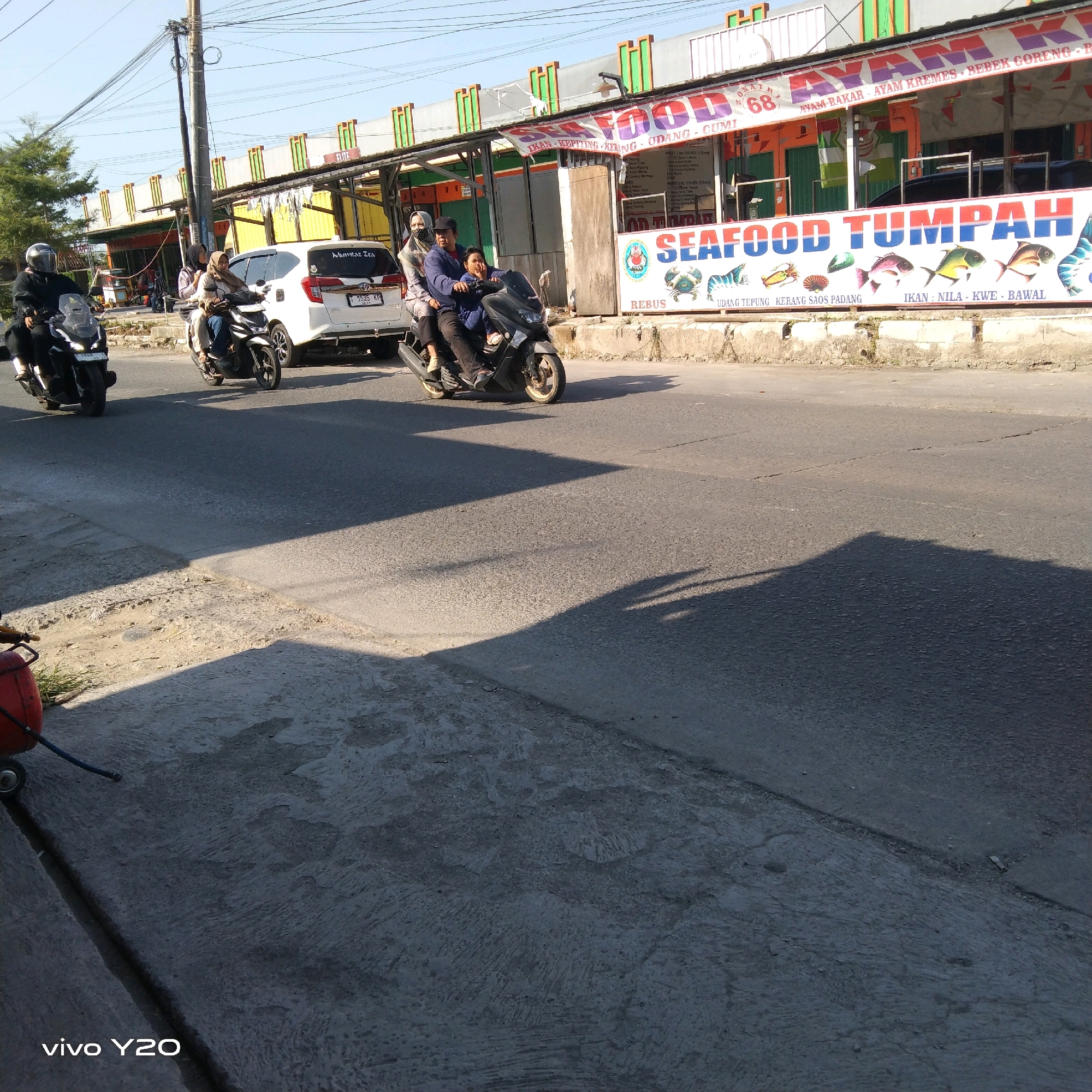 The image shows a street scene in an urban environment, likely in Southeast Asia based on the script on the signs. The foreground is a concrete sidewalk with a red object on wheels, possibly an air compressor or a fuel tank, with a hose. The main focus of the image is a road where several motorcycles and a white car are visible. On the left, a person wearing a black jacket and helmet is riding a black motorcycle. Further down the road, a family of three is on a black scooter: a man is driving, a woman is behind him, and a child is in front of the woman. A white car is parked to the right of the scooter. Behind the vehicles, there are buildings with colorful awnings and signs. One prominent sign has "SEAFOOD TUMPAH" written in large letters with illustrations of seafood and fish. Another sign above displays "AYAM BAKAR", "AYAM KREME", and "BEBEK GORENG". The environment appears to be a commercial area with open-air food stalls or restaurants. The lighting suggests it is daytime, with clear skies and visible shadows cast by the sun. The pavement on the road and sidewalk shows signs of wear and tear. The overall scene depicts a typical bustling street in a city, with everyday transportation and commercial activity.