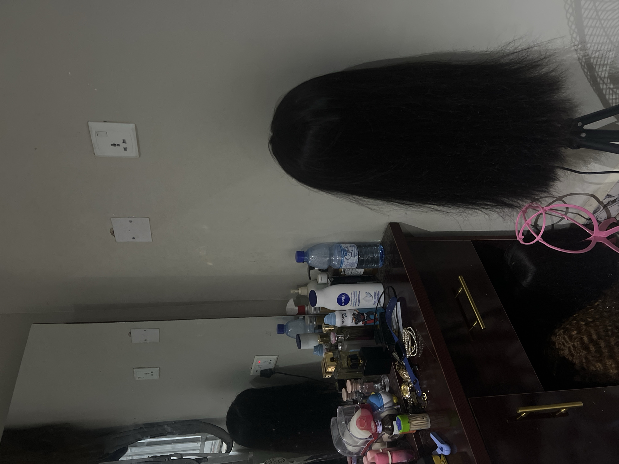 The image shows a vanity area in what appears to be a salon or personal grooming space in Howayi, Nigeria. A dark brown vanity table is positioned in front of a grey wall. On the table, there are numerous cosmetic and personal care items, including several bottles of perfumes and lotions, a bottle of Nivea body lotion, a bottle of water, and a container of toothpicks. A mirror is visible behind the vanity, reflecting a portion of the room. To the right of the vanity, a black wig is mounted on a stand. The wall has two electrical outlets. In the lower part of the image, a drawer is slightly ajar, revealing more wigs stored within. The overall impression is one of a functional, well-stocked grooming station.