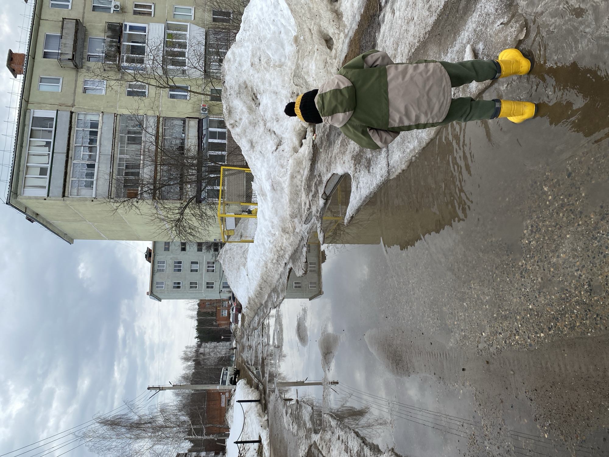 The photograph depicts a child standing in a puddle of water in Noviy, Russia. The child is wearing a green and beige jacket, green pants, a black and yellow hat, and yellow rubber boots. The child is standing next to a large pile of melting snow. The puddle reflects the image of the apartment buildings and the sky. The environment suggests it is spring time, with melting snow and bare tree branches. The buildings are typical Soviet-era apartment blocks. Power lines are visible in the background. The overall scene conveys a sense of early spring in a northern urban environment.
