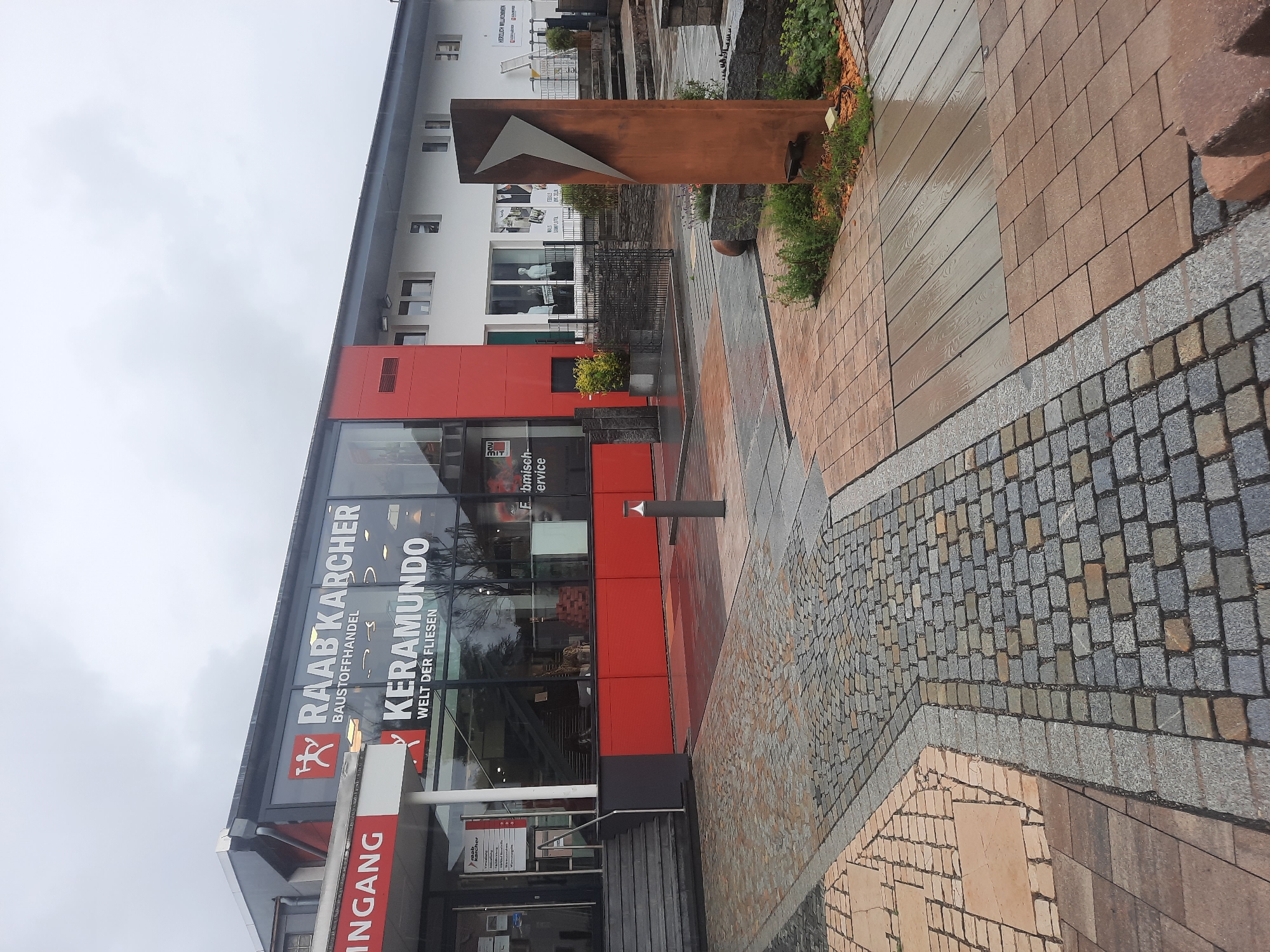 The image shows the exterior of a Raab Karcher building supply store, specifically the Raab Karcher Baumarkt with a Keramundo tile showroom, located in Rosenheim, Germany. The building features a modern design with large glass windows and a red accent facade. The entrance has a prominent "INGANG" sign. In front of the store, there is a display area with various paving stones and a metal sculpture. The ground is wet, suggesting recent rain. The sky is overcast. There are no people clearly visible in the image.