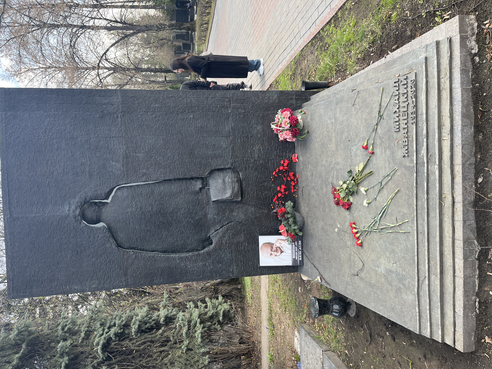 The image shows a tombstone in what appears to be a cemetery. The tombstone is made of dark, rough stone and features a silhouette of a man, likely Mikhail Zhvanetsky, whose name and life dates (1934-2020) are inscribed in Cyrillic on the adjacent slab. Flowers are placed on the grave, including bouquets and individual red and white carnations. A framed photograph of Zhvanetsky rests near the flowers. Two people stand near the tombstone, one of whom is observing it respectfully. The setting is outdoors, with trees and greenery visible in the background and a paved walkway nearby. The location is likely a cemetery in Moscow, Russia.