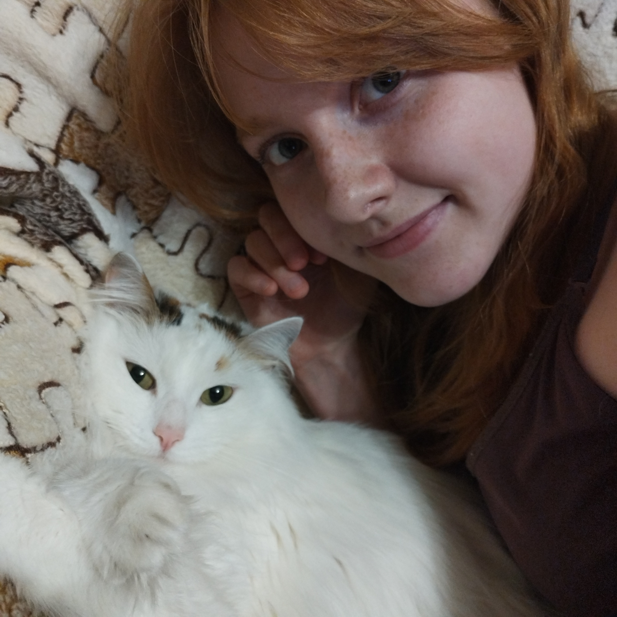 A young individual with red hair and freckles is seen lying down closely with a white, fluffy cat. The person, positioned in the upper right, has blue-grey eyes and a gentle smile, looking directly at the camera. Their red hair frames their face, and they are wearing a dark-colored top, possibly a tank top.
Beside them, occupying the lower left portion of the image, is a white cat with bright green eyes. The cat also appears to be looking towards the camera and has small patches of brown and orange fur on its head and ears. Both subjects are resting on a light-colored blanket or bedspread featuring a decorative brown pattern. The scene suggests a cozy, indoor setting, capturing a moment of companionship between the person and their pet. No visible text is present in the image.