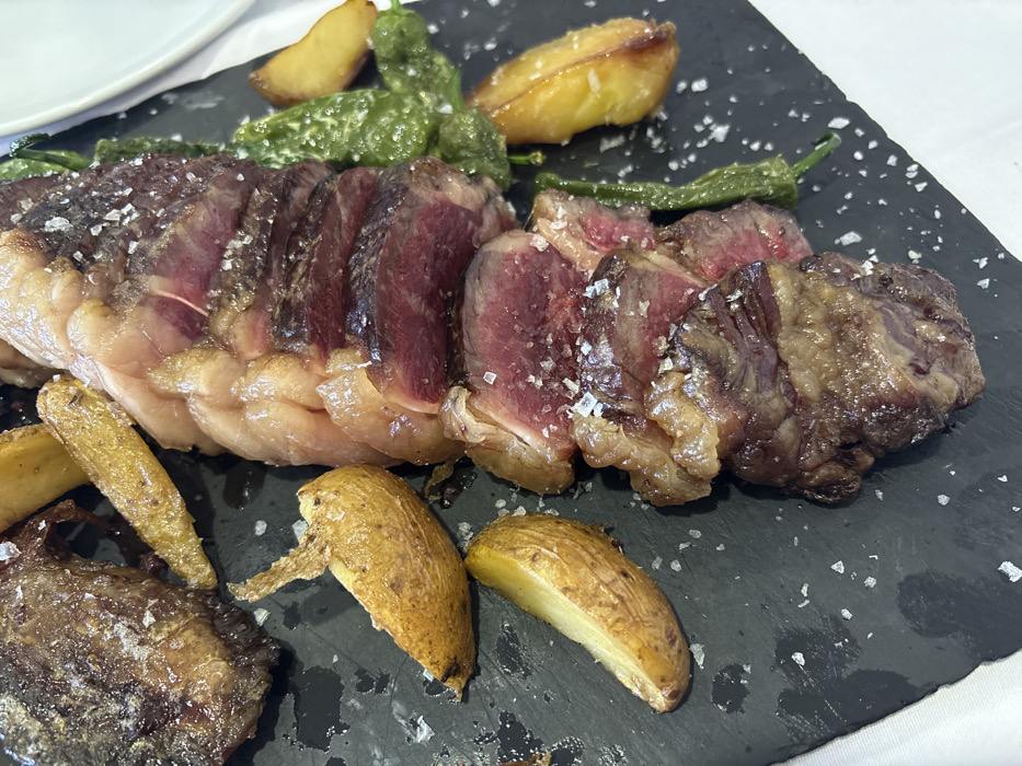 The image displays a close-up, overhead view of a meal served on a dark, rectangular, possibly slate plate, presented in a restaurant setting in Córdoba, Spain. The main subject is a large portion of sliced beef, exhibiting a rare to medium-rare doneness with a reddish-pink interior and a seared brown exterior. Notable rendered fat is visible along the edges of the meat slices. Coarse salt crystals are scattered across the meat and the plate surface. Accompanying the beef are multiple golden-brown potato wedges and several elongated green vegetables, consistent with peppers. A white tablecloth partially visible beneath the dark plate and a segment of a white round plate in the upper right background indicate a dining environment. No individuals are present in the frame.