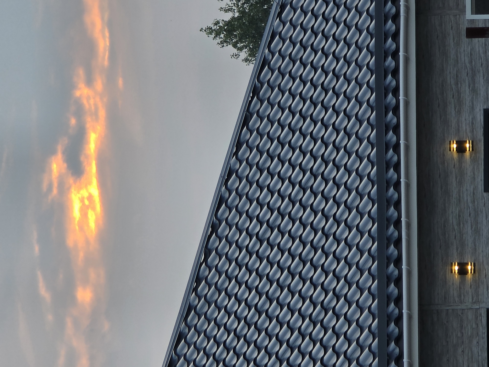 This is a low-angle, medium shot of a dark, wavy-patterned roof of a modern building. Above the roof, the sky is a soft gray with scattered clouds illuminated by a bright orange and yellow sunset. A few green leaves of a tree are visible in the upper right corner. The building's facade is visible below the roofline, constructed of grey, textured material, with two downward-facing exterior lights on either side of a central dark section. A white gutter runs along the edge of the roof above the facade. The overall scene suggests an exterior view of a residential or commercial building during dusk in Ермолаево, Russia.