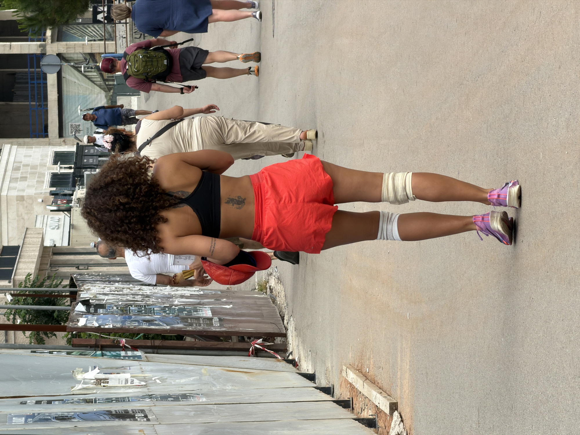 The image shows a sidewalk in Budva, Montenegro, where multiple people are walking. In the foreground, a woman with curly hair is wearing a black sports bra and red shorts. Her lower legs are wrapped in what appears to be white bandages or supports. She is holding a red baseball cap. Further up the sidewalk, there are a few other people walking, including a man with a backpack, a woman wearing light-colored pants, and a person in a blue shirt. A low wall covered with posters and graffiti borders the sidewalk on one side. On the other side, there is a glimpse of buildings, trees, and street signs. The street is paved and appears to be well-used.