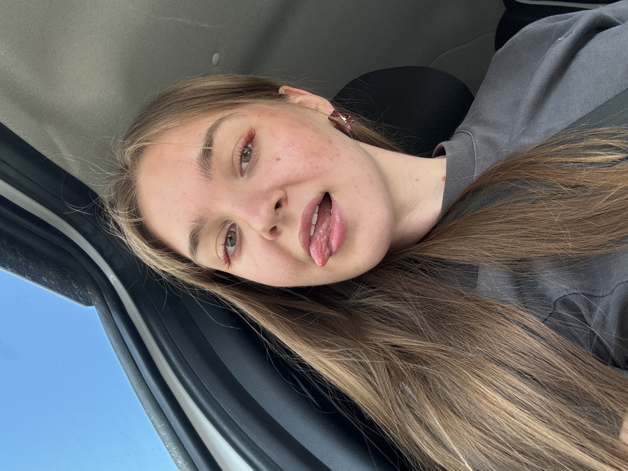 In a low-angle, close-up selfie, a young woman with long, light brown hair is seated in a vehicle. She is sticking her tongue out playfully, with her mouth slightly open. Her eyes are a light green, and she is wearing bright red eyeshadow and matching earrings. Her skin appears fair with some light freckles and blemishes. She is wearing a dark gray t-shirt. The interior of the vehicle is visible, with the black trim of a car door to her left and a hint of the car's headliner and a bright blue sky through an opening above. The overall impression is a casual, indoor shot, likely taken from the passenger seat of a car. There are no identifiable landmarks or features that would place this image in Perm, Russia or any other specific city.