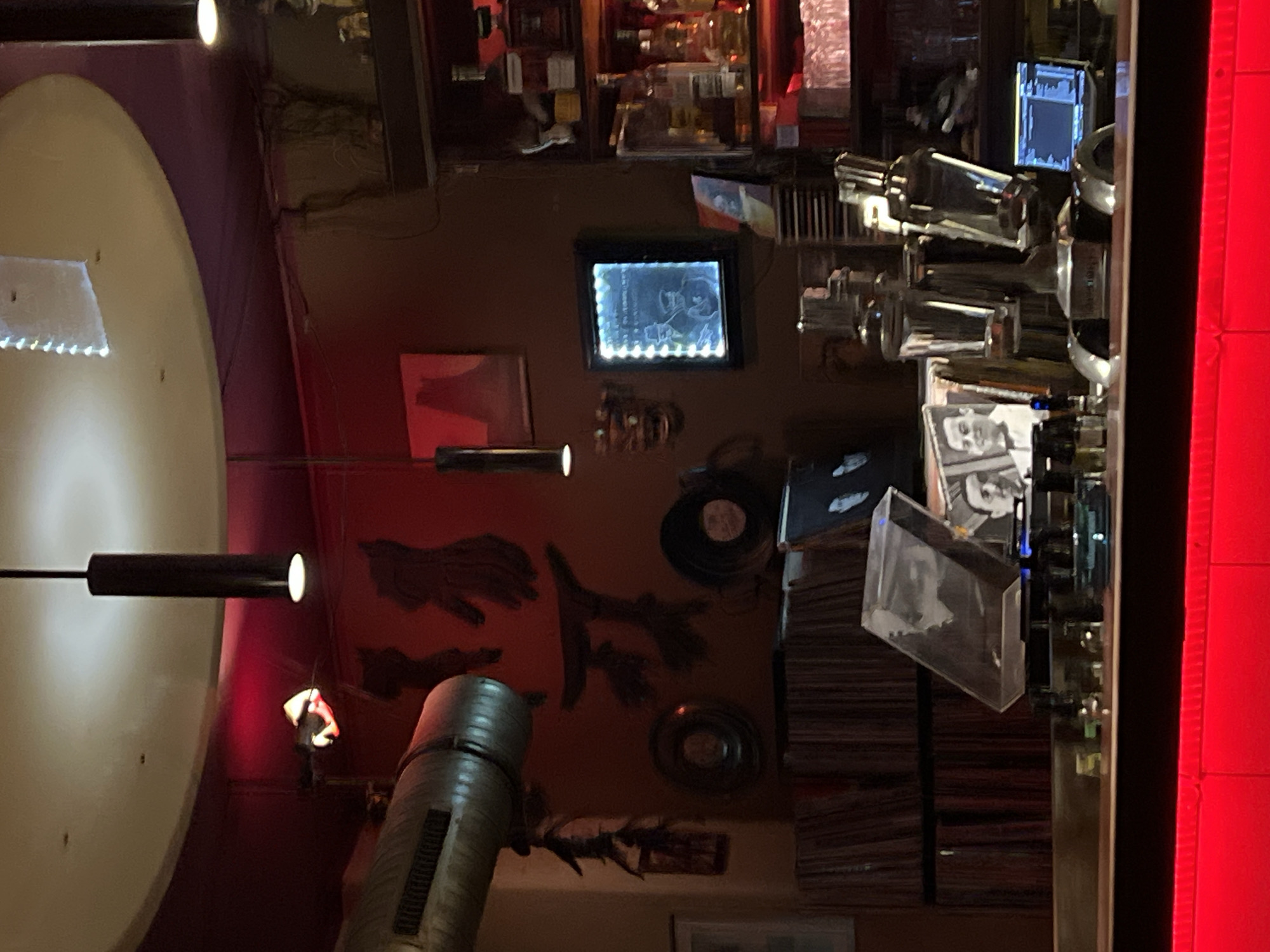 The image shows an interior scene of a bar or club with various elements suggesting a retro or music-themed establishment. There are lights, framed images on the wall, records and record cases stacked on a shelf, and bar equipment like drink dispensers. The ambiance is dark with accents of red light. It may be a bar located in the city of Berlin, Germany.
