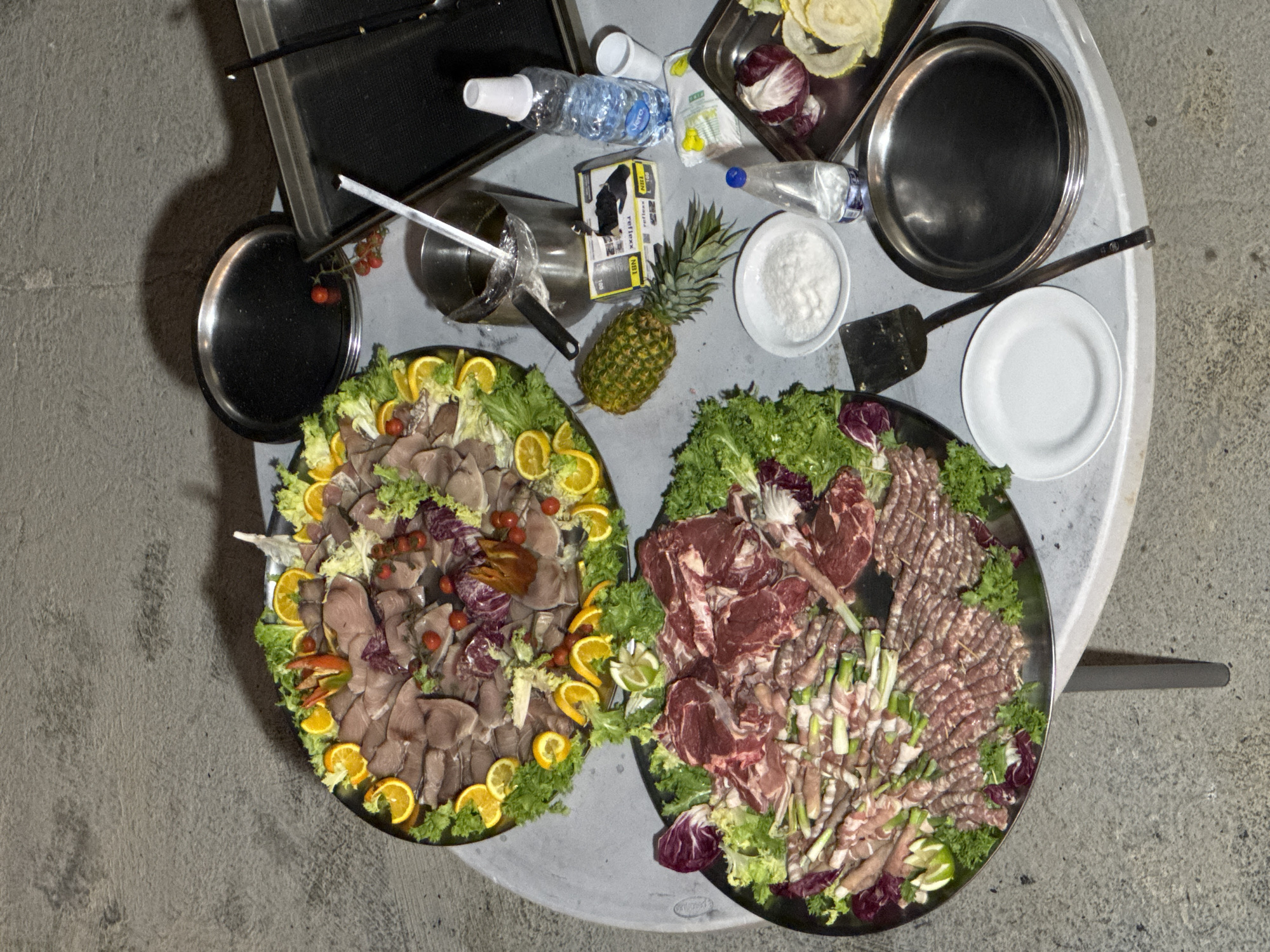 An overhead, medium shot captures a rustic outdoor table laden with a variety of food items. The table, a light gray, round surface with a slightly textured finish, is positioned on a concrete ground. To the left, two large, dark platters are filled with food. The upper platter showcases thinly sliced, cooked meats, possibly fish or cured meat, arranged over a bed of lettuce and garnished with orange slices, cherry tomatoes, and red radicchio. The lower platter presents raw cuts of meat, including what appear to be steaks and rolled meat skewers with green onions, also nestled on lettuce and radicchio. To the right of the platters, several items are scattered across the table. A whole pineapple with its spiky green top sits prominently. Adjacent to it, a stainless steel pot with a long handle contains a liquid, with a spatula resting inside. Next to the pot, a clear plastic bottle of water stands upright. A small box, labeled in black text, is also visible. Further to the right, a stack of empty, shiny metal plates and a white plate rest on the table. Another clear plastic bottle, this one with a blue cap, is present. In the background, a dark, rectangular serving tray and a black circular plate are partially visible. The scene is lit from above, casting shadows on the concrete floor and the objects on the table. The overall impression is that of a prepared meal, perhaps for an outdoor gathering.