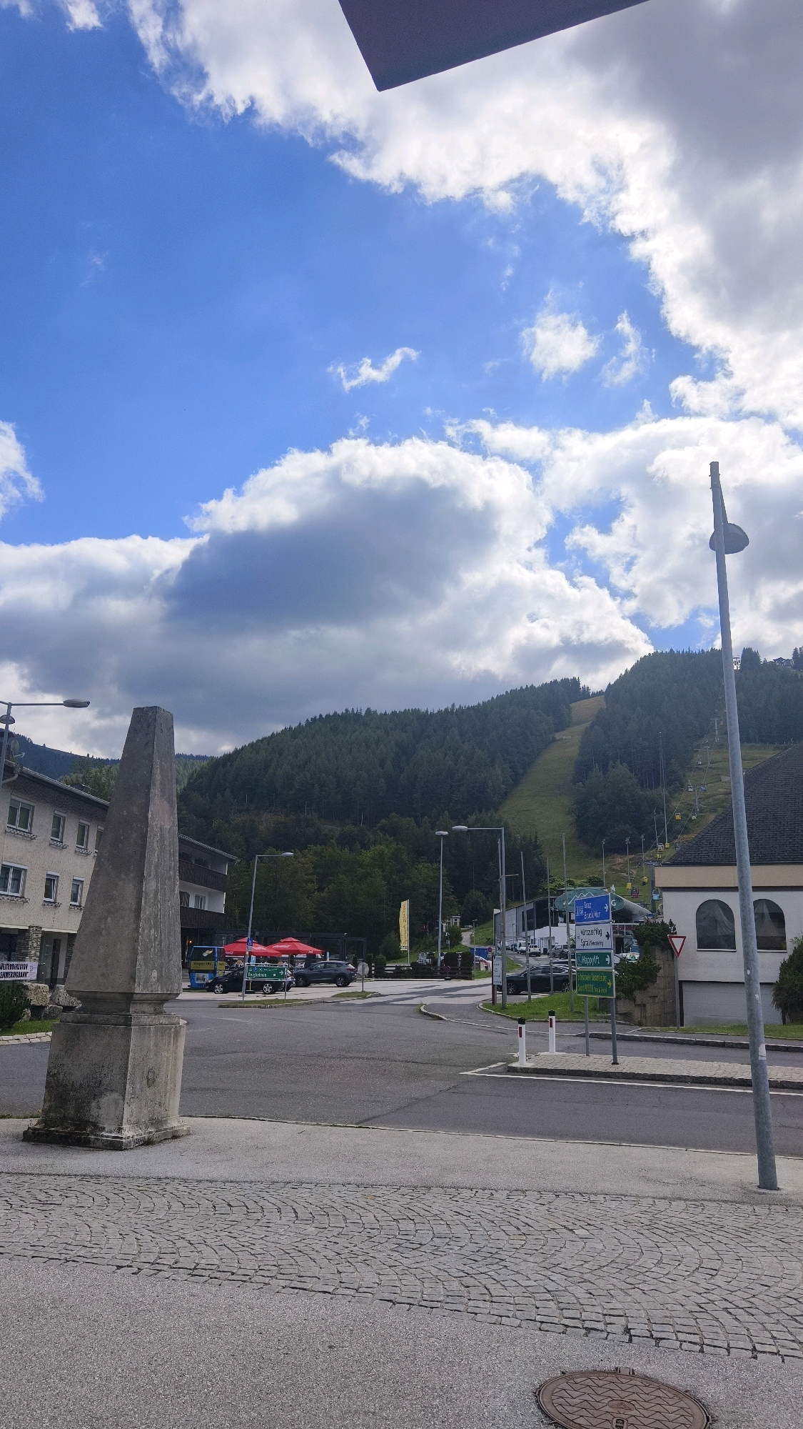 The image shows a street scene in Spital am Semmering, Austria. In the foreground, a cobblestone pavement leads to an asphalt road. To the left, a tall, weathered stone monument stands prominently. Several buildings are visible on the left side of the street, one of which has a sign with German text. On the right side, a light pole with a streetlamp is present. Further down the street, a ski slope is visible on a forested mountain, with ski lifts ascending the slope. Various vehicles, including cars and a bus, are parked or driving on the road. Several traffic signs are also visible, providing directional information. The sky above is blue with scattered white clouds, suggesting a clear day. Red umbrellas are set up outside a building on the left, possibly indicating an outdoor seating area for a cafe or restaurant. The overall impression is a quiet, possibly rural, Austrian town with a strong connection to its mountainous surroundings.