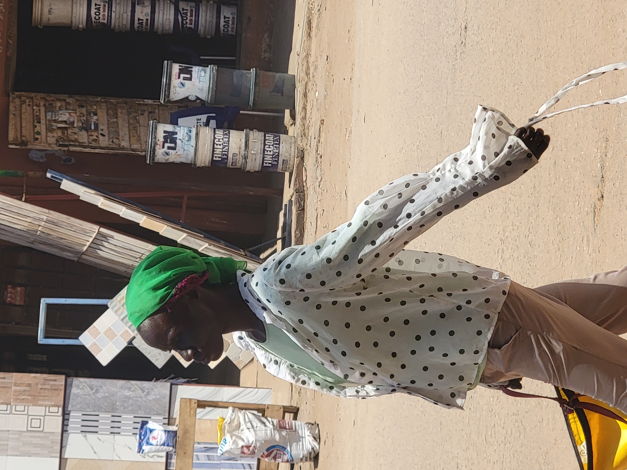A person wearing a green head covering, a white shirt with black dots, and tan pants stands outdoors in what is likely Maiburiji, Nigeria. The person is facing left and has their right arm raised, holding white straps. In the background, there is a storefront with stacked paint buckets and various tiles displayed. A yellow bag is visible near the person's lower body. The ground is light brown, likely a street or sidewalk.