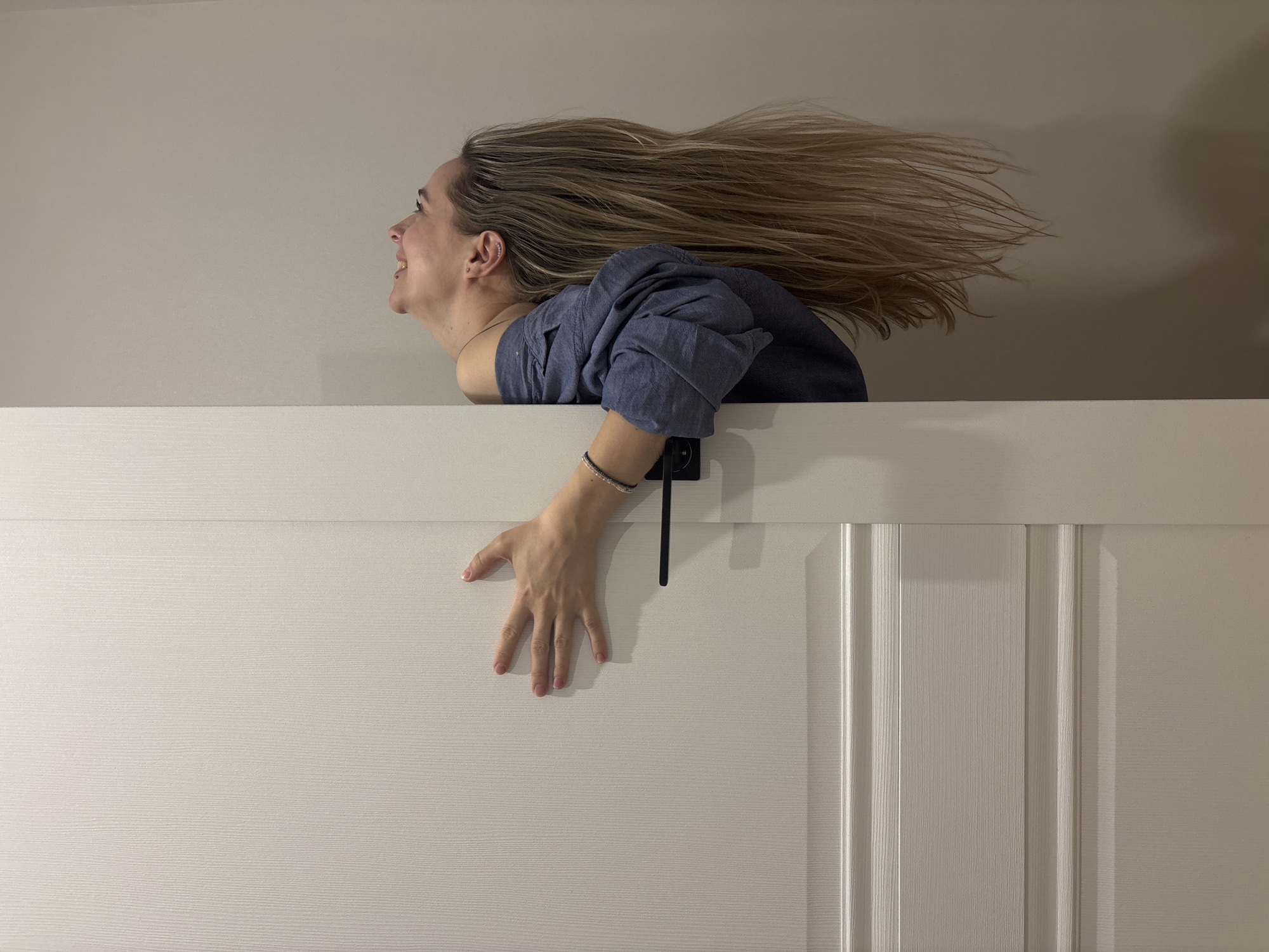 A woman with light brown hair and wearing a blue shirt is positioned above a white paneled door in what appears to be an indoor setting. The woman is looking upwards and smiling, with her hair spread out in the air above her. Her hand is reaching down onto the door. The background is a plain, light beige wall. There is no indication of the location being Chaikovsky, Russia, from the image alone.