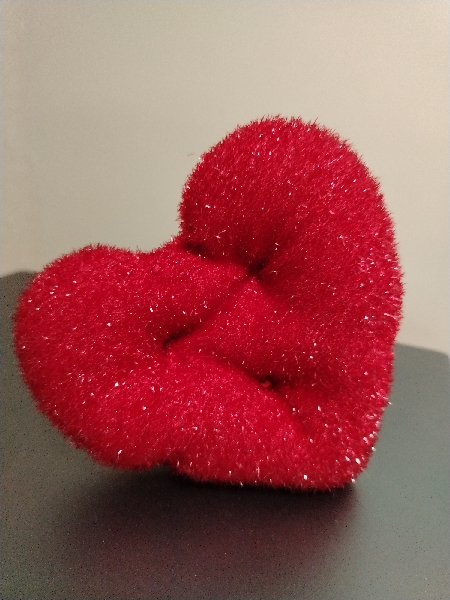 A fluffy, red, heart-shaped object is positioned on a dark, flat surface. The object has a textured, sparkly surface. The background is a plain, light-colored wall. The image appears to be a close-up shot of the object. There is no information about its location in Krasnodar, Russia, as this is not discernible from the visual content of the image.