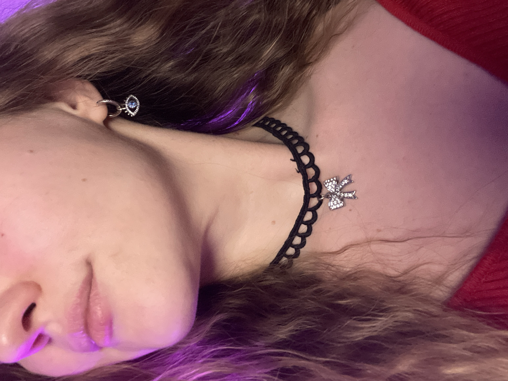 A young person with fair skin is pictured from the side, lying down with their head tilted slightly. They have brown hair with a strand dyed purple near the face. The person is wearing a black lace choker with a silver butterfly pendant, and a silver earring with a blue gem. A red garment is visible at the top of the frame. The setting is undefined and the image contains no visual cues as to being in Batumi, Georgia.