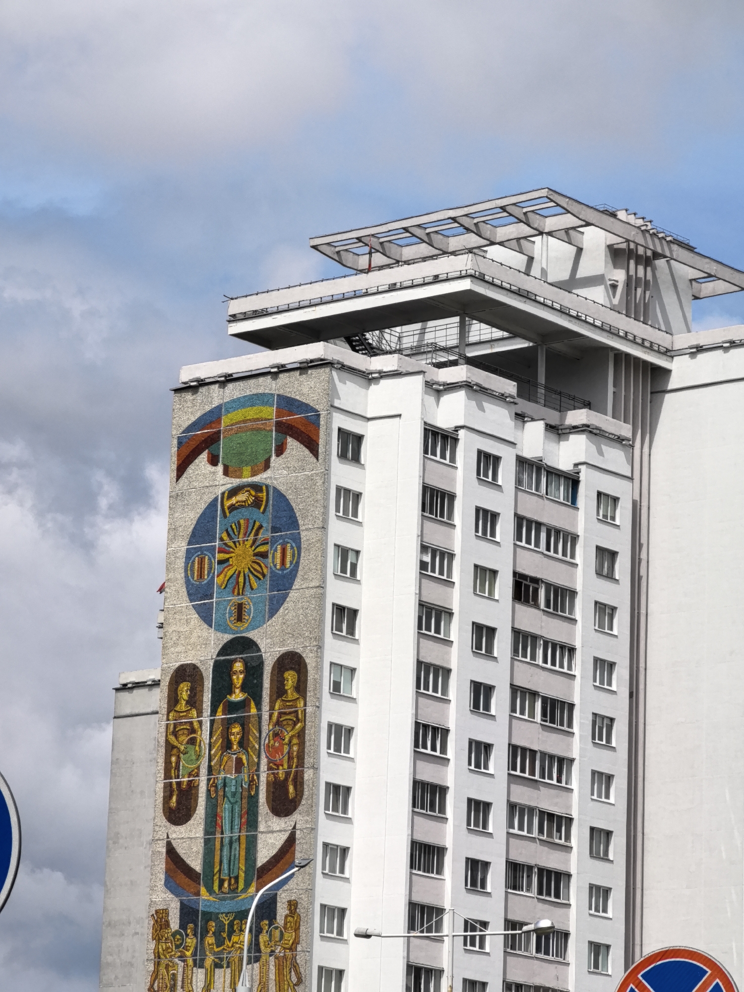 The image shows the exterior of a tall, multi-story building in an urban environment. The building is primarily white and gray with numerous windows. A large, colorful mosaic mural is featured on the left side of the building's facade. The mosaic depicts various figures and symbols, including a sun-like emblem, a rainbow, and groups of people. Above the main residential floors, there is a distinct architectural element with a geometric, open-frame design. The sky above is cloudy, with patches of blue visible. In the foreground, a portion of a blue and red circular traffic sign is visible on the left, and another partially visible red and blue circular sign with a diagonal line is on the lower right. A street light is also present in the lower part of the image, in front of the building. The overall impression is of a modern, somewhat brutalist architectural style, accented by the vibrant mosaic artwork.