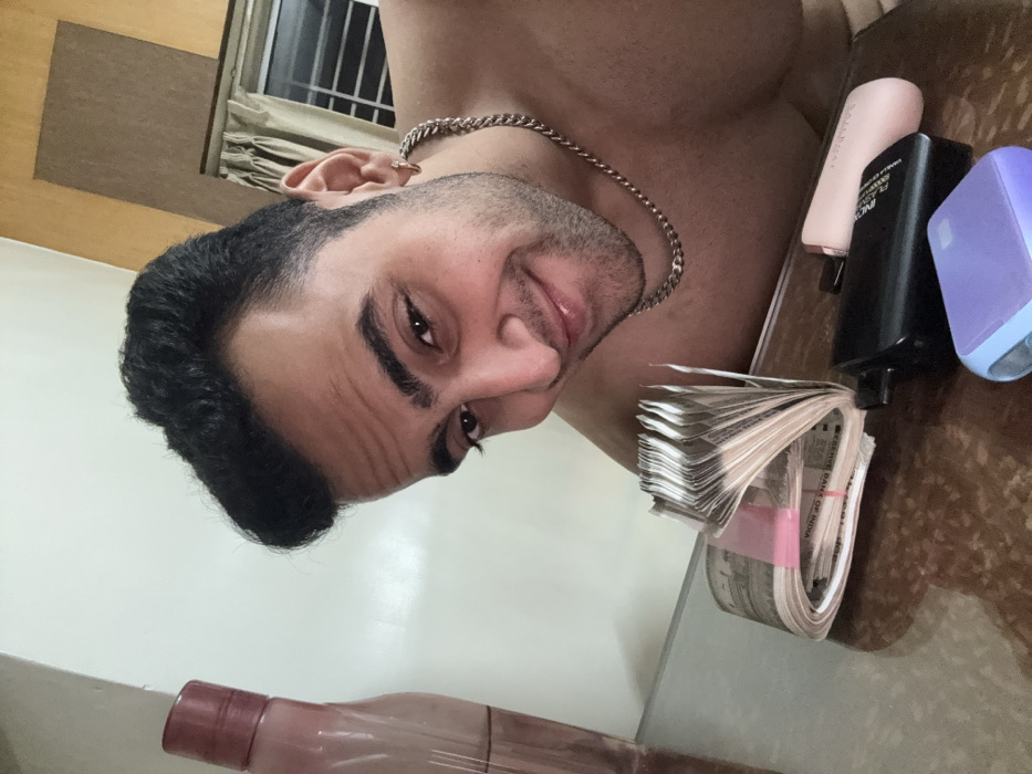 A man poses close to the camera, revealing his chest and a chain necklace. He is partially shown with a smirk. Stacked Indian Rupees are in focus. Beside the money, there are cosmetics, a black power bank, and a blue item. A water bottle is present in the lower portion of the image. A window can be spotted in the top portion of the image.