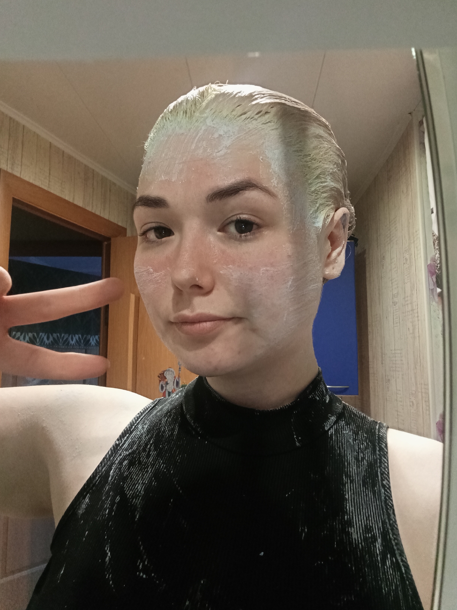 A medium shot captures a young person taking a mirror selfie indoors. The subject, with fair skin, dark eyebrows, and brown eyes, looks directly at the camera with a calm, neutral expression, possibly a slight smile. Their short hair is covered in a light, possibly pale green-tinged, creamy substance, suggesting a hair treatment like bleach, dye, or toner. White streaks of the same substance are visible on their cheeks and forehead. They are wearing a black, ribbed, high-neck sleeveless top, which also has streaks of the white substance on it. A hand extends from the left, forming a peace sign with two fingers.
The setting appears to be a room, likely a bathroom, with light-colored wooden paneling on the walls. A wooden door frame is visible on the left, leading to a space with a prominent dark blue wall or object. The ceiling is light-colored with decorative molding. On the wall behind the subject's right shoulder, some small, colorful figures or drawings can be vaguely discerned. The image is a reflection, with the edge of a mirror frame subtly visible on the right side of the photo. No text is visible in the image.