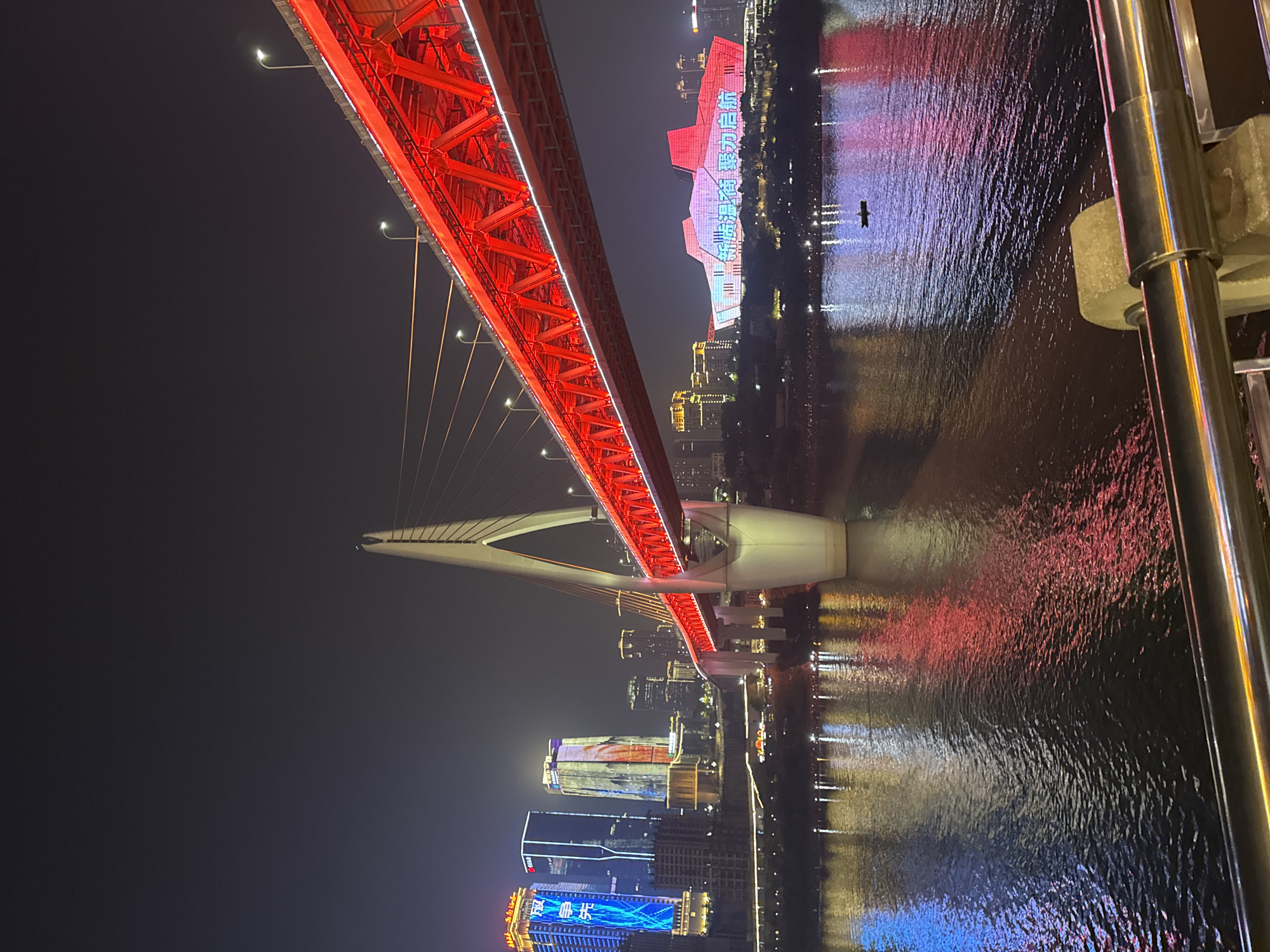 At night, a red suspension bridge spans a body of water, likely the Yangtze River, in the Yuzhong District of Chongqing, China. The bridge is lit with white lights. A distinctive building, also brightly lit with red, white, and blue, reflects on the water. High-rise buildings with illuminated signs are visible in the distance. A black object, possibly a boat, appears on the water, and a metal railing is visible in the foreground.