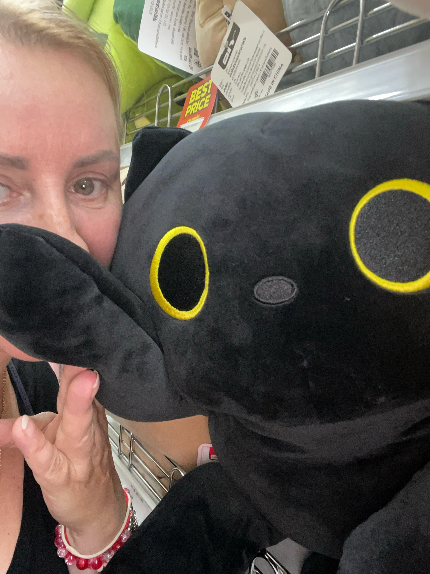 A close-up selfie shows a woman's face and eye peering from behind a large black plush toy with yellow circular eyes. The woman has blonde hair and is wearing a black top and a red and white beaded bracelet on her left wrist. In the background, shelves are visible with merchandise, including what appears to be stuffed animals. A "BEST PRICE" tag is also visible. The scene appears to be a retail store, likely in Pattaya City, Thailand, given the presence of Thai text on some of the labels. The toy is positioned to obscure a significant portion of the woman's face and the background.