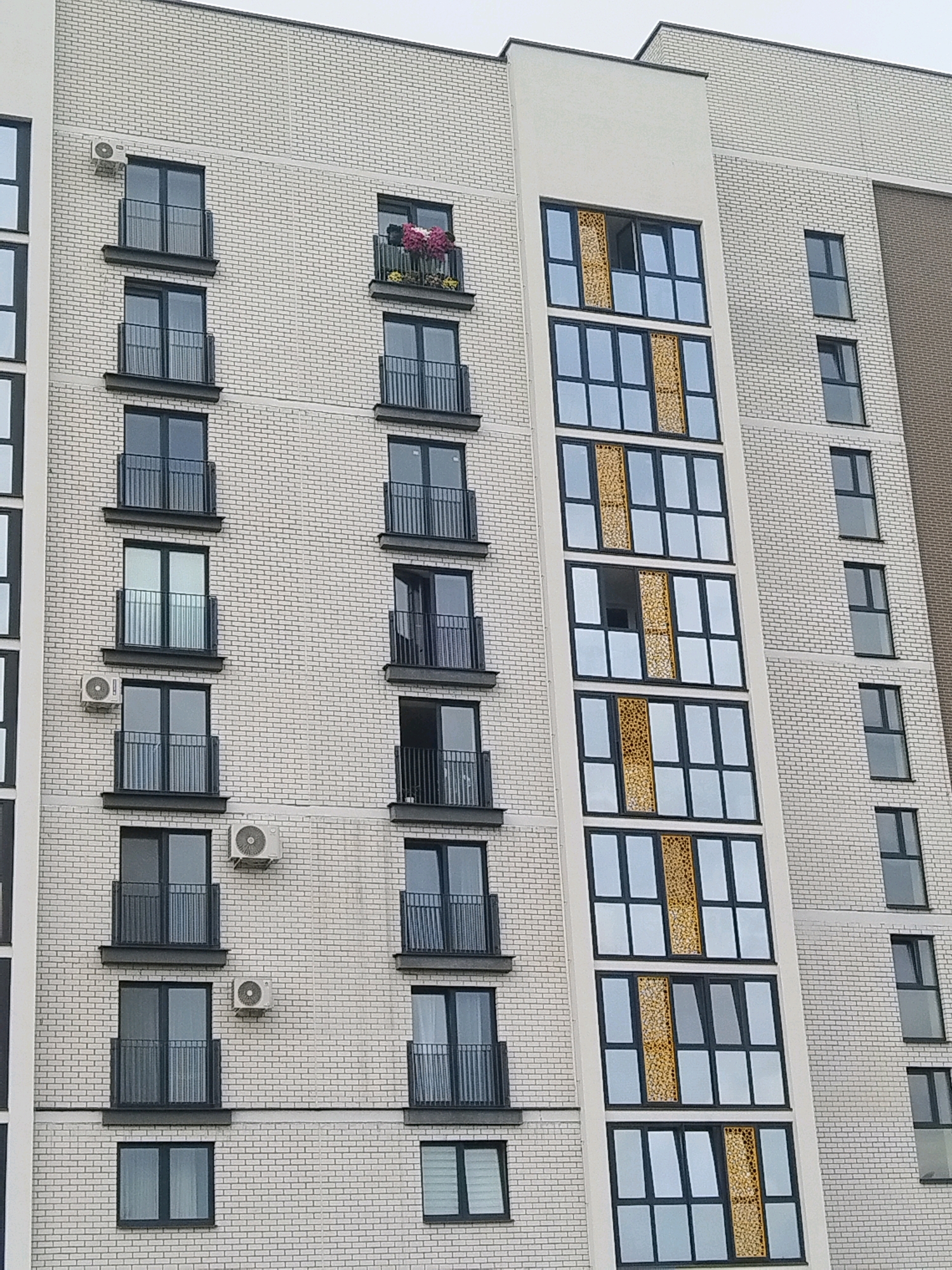 The image shows a modern apartment building with a light brick facade. The building has multiple stories, each with numerous windows and balconies. Some balconies contain flowers. There are also air conditioning units visible on the exterior walls. The building's design includes sections with large, multi-paned windows featuring decorative golden panels. The overall scene depicts a residential area in the city of Brest, Belarus.