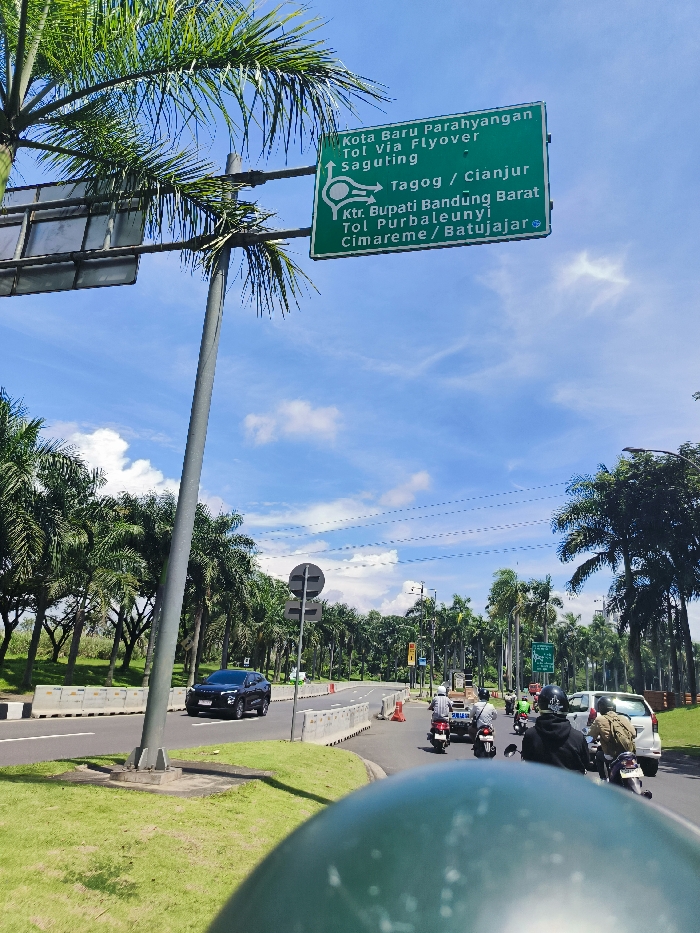 The image shows a street in Padalarang, Indonesia (latitude: -6.852, longitude: 107.495). A green street sign with white lettering indicating directions to various locations, including Kota Baru Parahyangan, Tol via Flyover Saguting, Tagog, Cianjur, Ktr. Bupati Bandung Barat, Tol Purbaleunyi, Cimareme, and Batujajar, is mounted on a pole at the top of the image. The pole is tilted to the right and is set on a grassy median between a paved road and a sidewalk. A black car is driving on the paved road, with green vegetation and several palm trees lining both sides of the road in the distance. A group of people is riding motorcycles on the paved road in the distance. The sky is blue with a few white clouds. The image is taken from the perspective of a person sitting on a motorcycle. The motorcycle's seat is blurred, suggesting that the person taking the image is on the motorcycle.
