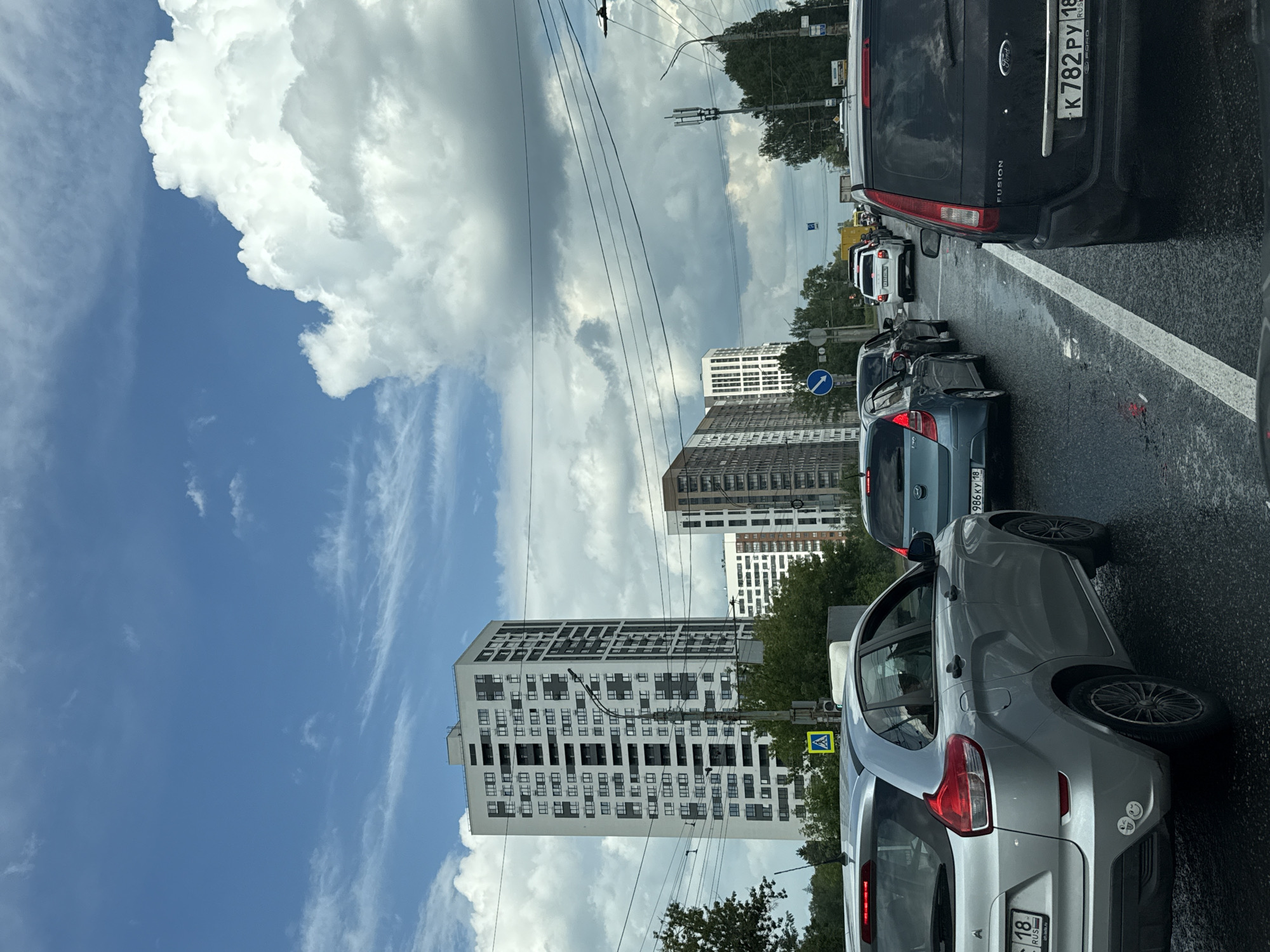 This image depicts a traffic jam on a city street. In the foreground, a silver hatchback is visible, with its rear quarter panel and bumper taking up the left side of the frame. A black SUV is positioned to the right of the silver car, with its rear bumper and license plate clearly in view. Several other vehicles are lined up behind them, suggesting a significant traffic congestion.
The street appears wet, indicating recent rain or damp conditions. The environment is urban, with several tall, modern buildings lining the right side of the street. One building is particularly prominent, a tall, multi-story structure with a white facade and numerous dark-colored windows. In the background, more buildings and trees are visible, along with power lines stretching across the sky. The sky is partially cloudy, with large white cumulus clouds against a blue backdrop. A yellow pedestrian crossing sign is also visible near the silver car.
The license plates on the vehicles suggest the location is Russia, specifically the city of Izhevsk. The silver car has a license plate starting with "18", which corresponds to the Udmurt Republic, where Izhevsk is the capital. The black SUV has a license plate starting with "K 782 PY", also a Russian format. The presence of multiple modern high-rise buildings and the specific license plate details strongly suggest this scene is in Izhevsk, Russia.