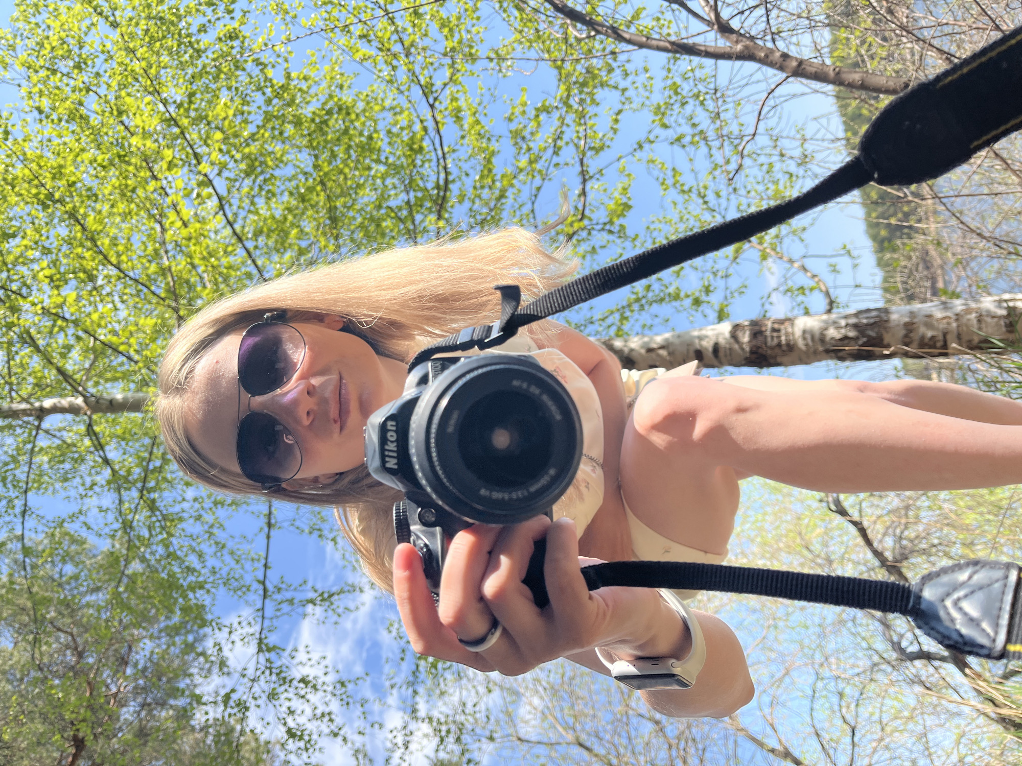 A woman holds a Nikon camera, facing the lens, standing outdoors amidst trees. She wears sunglasses, a watch, and a ring. The background includes blue sky and green foliage. The city of Chaikovsky, Russia, is not discernible in the provided image.