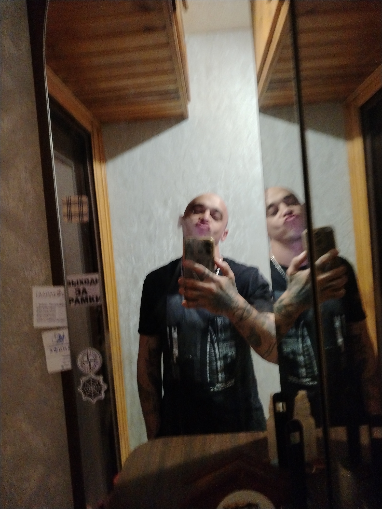 A bald man with tattoos on his arms takes a selfie in a mirror. He is wearing a black t-shirt and is puckering his lips, appearing to blow a kiss or smoke. The mirror reflects another image of the man and a portion of the room. The room appears to be a bathroom or dressing area, with wood-paneled cabinets above the mirror and textured walls. There are several stickers on the wall to the left of the mirror, including one with Russian text that appears to say "выход за рамки" (outside the box). The lighting is dim.