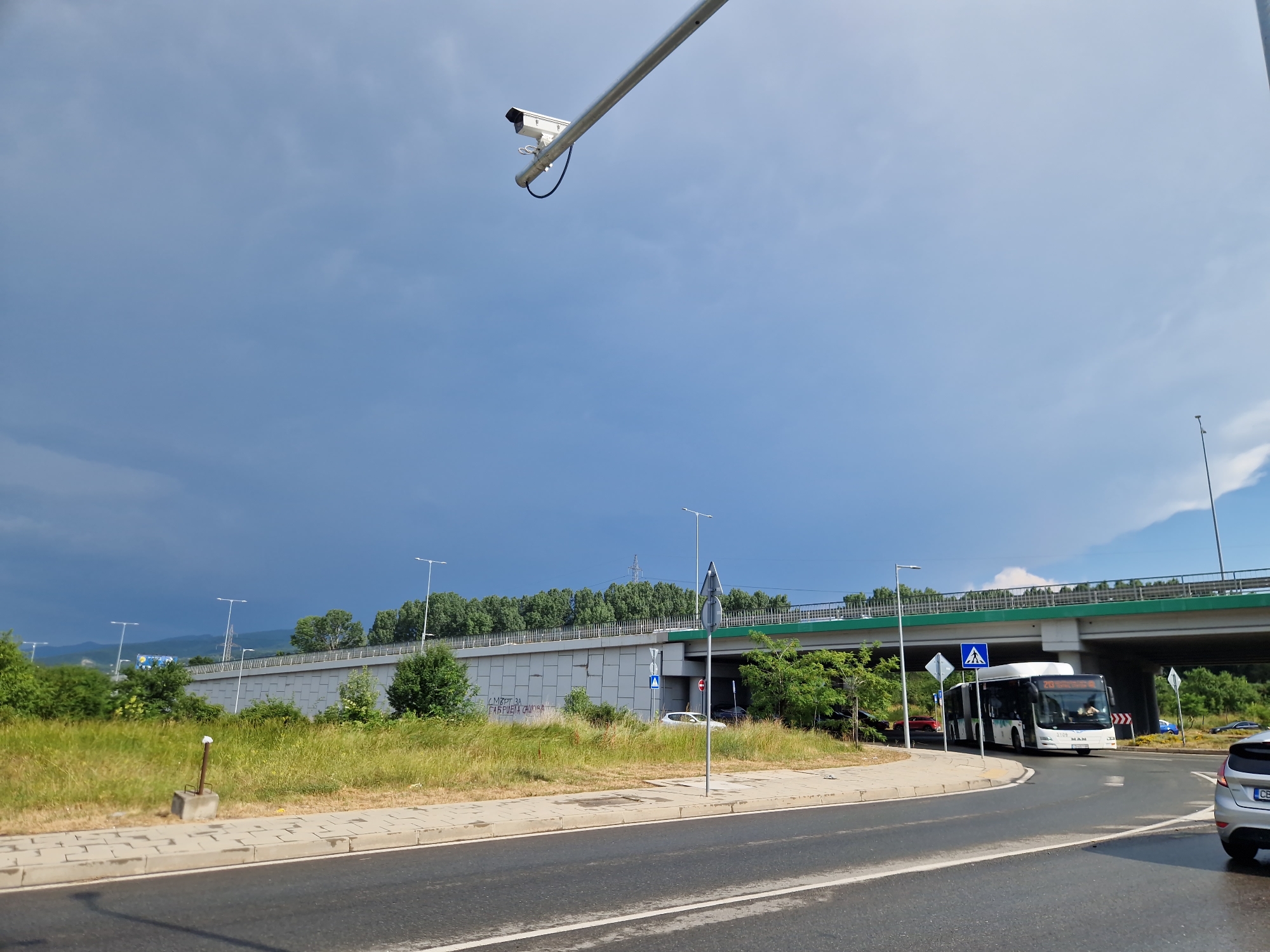 The image depicts a street scene in Sofia, Bulgaria, under a partly cloudy sky. The weather appears to be transitioning, with dark, ominous clouds gathering in the distance, suggesting an impending storm, while patches of blue sky are visible. The time of day is likely afternoon, given the lighting.
The foreground shows a paved road with lane markings and a sidewalk. On the sidewalk, a small concrete base with a metal pole protrudes from the ground. To the left, a patch of overgrown grass and vegetation lines the roadside.
In the midground, a large concrete wall, possibly part of an overpass or sound barrier, runs horizontally across the scene. Grafitti is visible on the wall. Beyond the wall, a lush green belt of trees and foliage is present.
A modern overpass with a green underside is prominent, spanning across the scene. Streetlights are visible along the overpass and on the side of the road.
A white bus with the number "28" displayed on its front is turning onto a road under the overpass. The bus has "MAN" written on its front, indicating the manufacturer. Several other vehicles, including a white car and a red car, are also visible in the vicinity of the overpass. A silver car is parked on the right side of the image, with a license plate partially visible.
A security camera is mounted on a pole extending from a metal arm above the road, pointing towards the street. The overall scene suggests an urban or suburban environment with significant infrastructure. The graffiti on the wall appears to be in Cyrillic script, consistent with the location in Bulgaria.