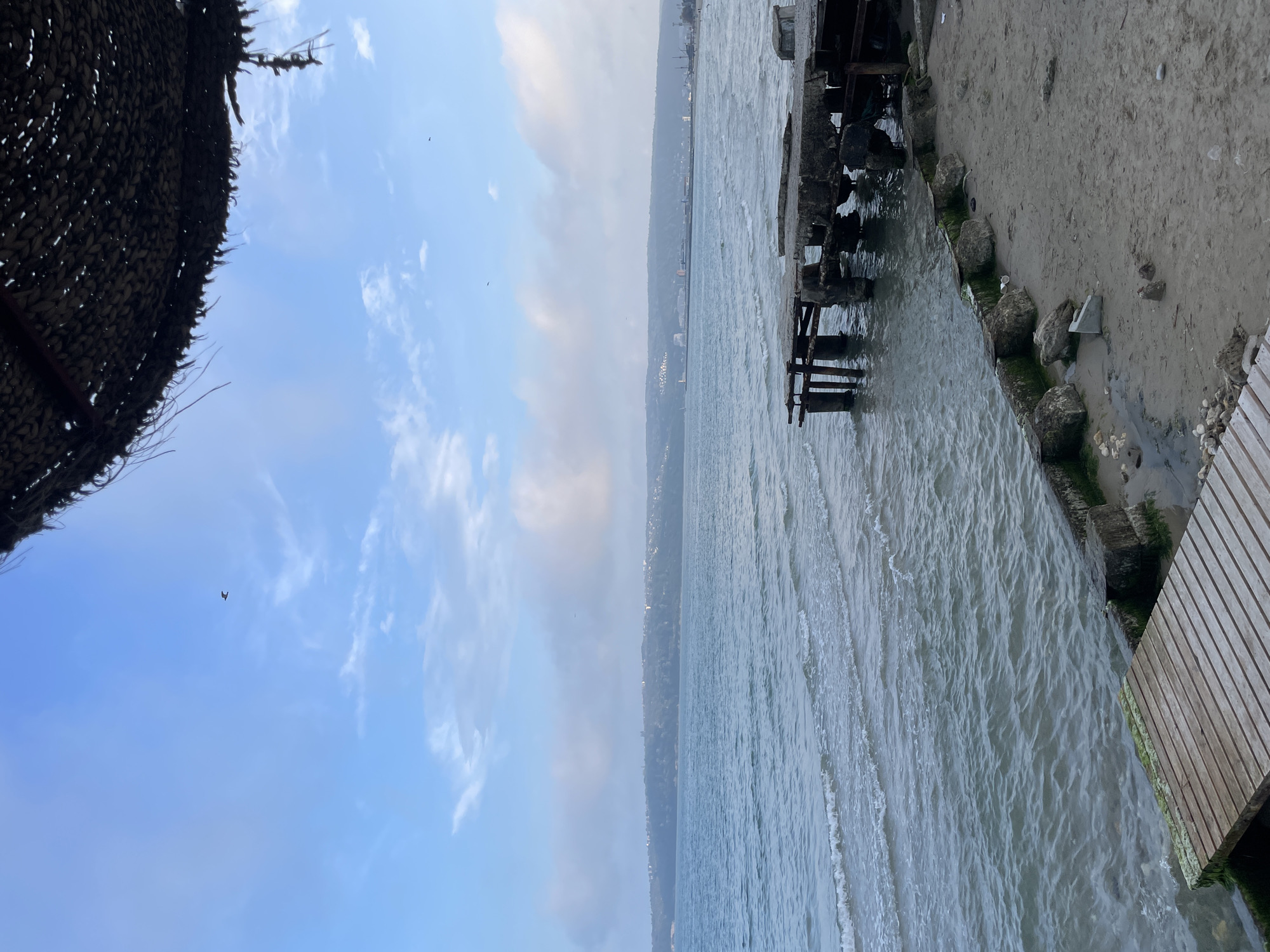 The image shows a view over the Black Sea from Varna, Bulgaria, with a weathered pier structure in the foreground. The pier's construction includes concrete, timber, and stone, showing signs of age and possible disrepair. The waterline reaches the pier's base and a small beach area. The sea is choppy, with visible wave action. The horizon shows a hazy cityscape. To the left, a thatched structure partially obscures the sky, which is light blue with some cloud cover. A bird is visible in the sky.