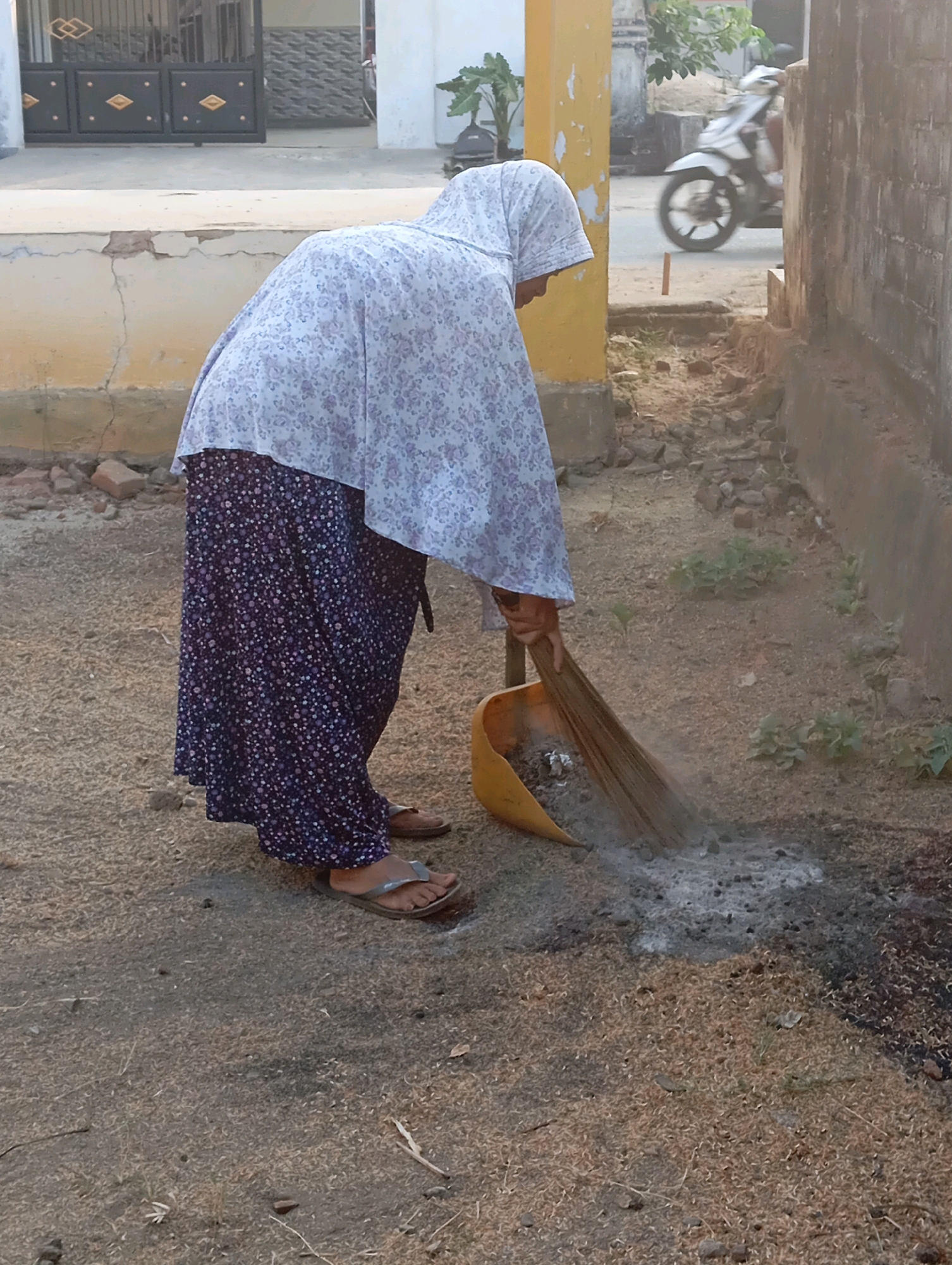 A person wearing a floral hijab and a long floral skirt is sweeping debris with a broom into a yellow dustpan on a dirt path. The person is bent over, facing away from the camera. In the background, a white motorcycle is visible on a road, and parts of buildings and a fence can be seen. The scene appears to be in a residential area of Adi Puro, Indonesia.
