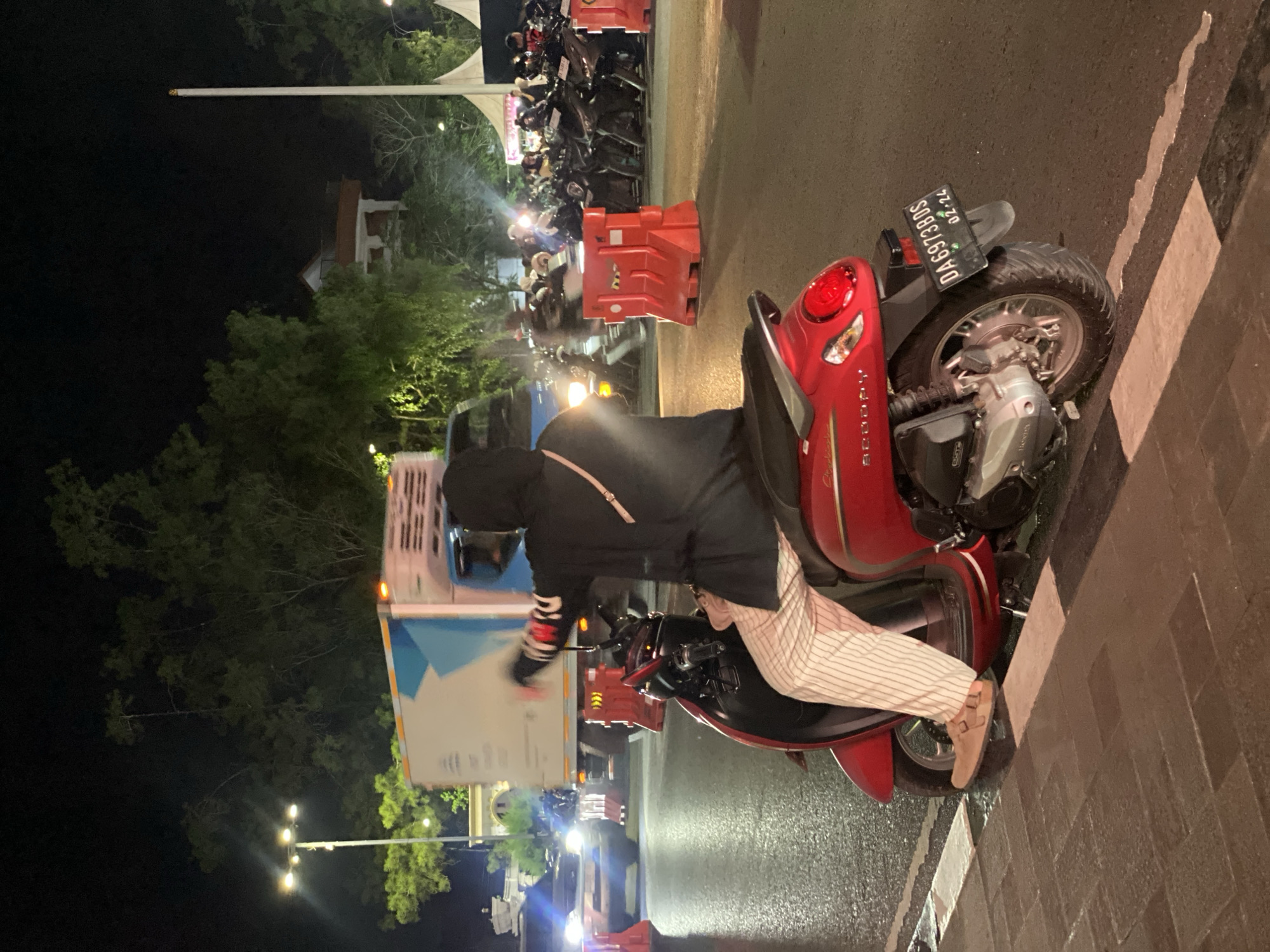 A person wearing a black hoodie and striped pants sits on a red scooter at night in Banjarbaru, Indonesia. The scooter has a license plate that reads "DA6973BDS 02-24". The person is looking to the right and their left arm is raised. In the background, a large white truck is driving past, and many other scooters are parked in a lot. There are red traffic barriers and trees visible. The ground is wet, indicating it may have recently rained.