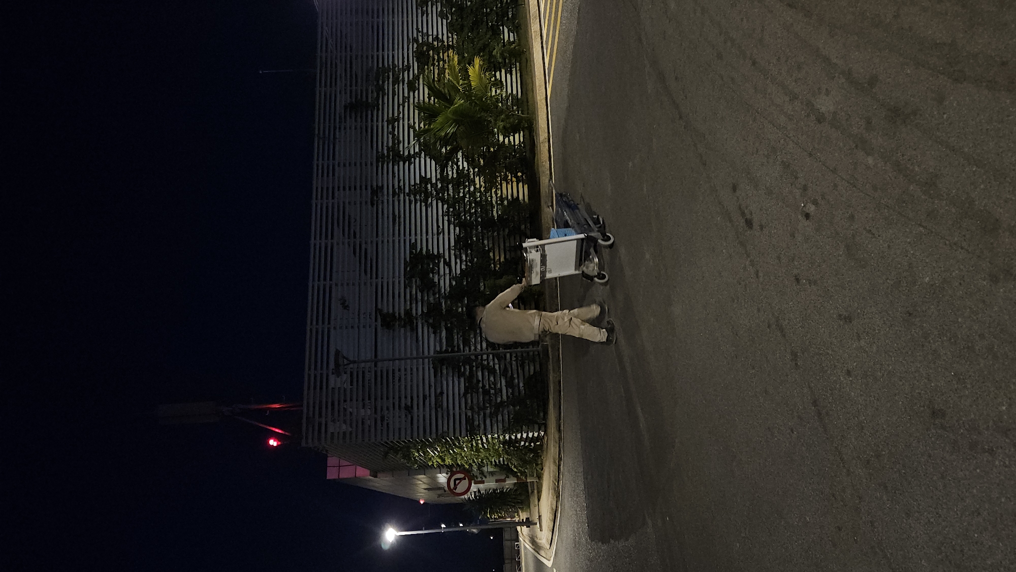 The image captures a nighttime scene in Cam Ranh, Vietnam, showing a solitary figure pushing a utility cart along a road beside a modern building. The scene is illuminated by various artificial light sources, casting shadows and highlighting specific elements.
In the foreground, a person, likely male, is dressed in light-colored, possibly beige or tan, long-sleeved clothing and trousers, with dark footwear. They are actively pushing or guiding a white, two-wheeled utility cart or trolley. A blue item or bag is placed on the lower section of the cart.
To the left of the person, a tall, contemporary building features a prominent slatted or louvred facade, appearing light in color under the ambient lighting. In front of this building and along the roadside, lush tropical vegetation, including palm trees with bright green fronds and dense bushes, adds a natural element to the urban setting.
The person and cart are on a dark asphalt road surface that shows textural details and subtle reflections from the scattered lights. A yellow line is visible on the edge of the road. Further down the street, a street light casts a gentle glow. Near this light, a red circular "no U-turn" sign is visible, displaying a curved arrow within a red circle and slash. High in the dark night sky, in the distance, a distinct red light, possibly a warning light or beacon, is also visible. The overall atmosphere is quiet and somewhat serene, indicative of late hours.
