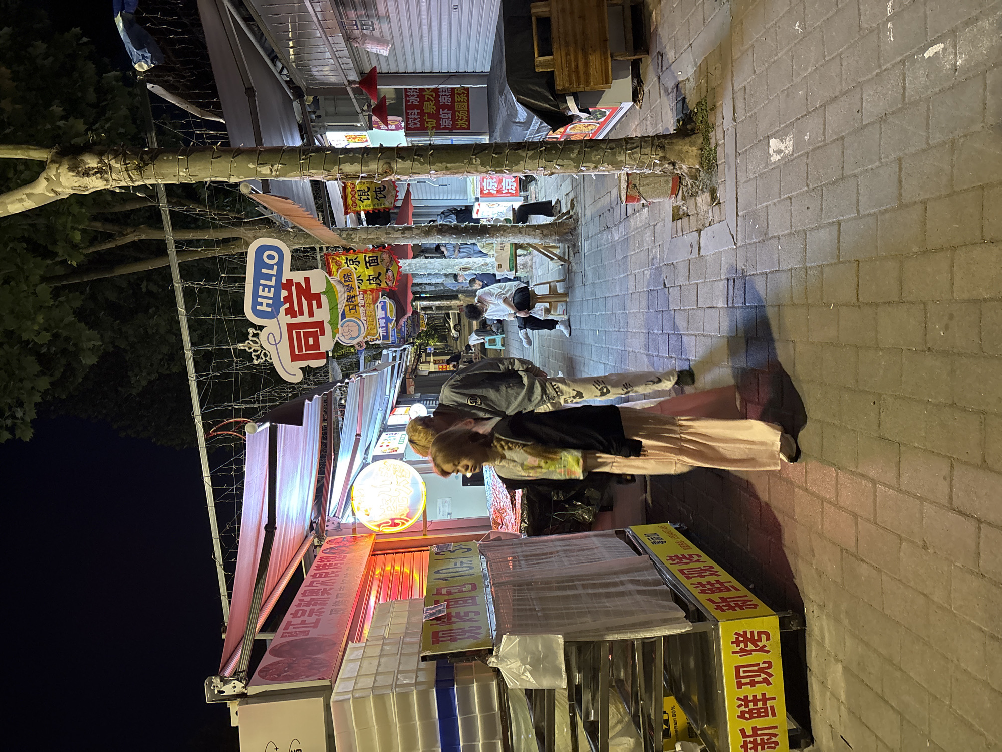 Here is a detailed description of the image:
**Overall Scene:** The image depicts an outdoor food stall scene in Jiangjin, China, at night. The stalls line a paved sidewalk.
**People:** There are several people visible. A couple is standing at the front of the stall, seemingly interacting with it. Other individuals are sitting on chairs further down the sidewalk, potentially customers at other stalls.
**Objects:**
* **Food Stalls:** The stalls are covered with awnings and display various food items and signs written in Chinese. The closest stall has lighted signs with Chinese text.
* **Signs:** There are several signs visible. One sign has "HELLO" written in English. Several others are written in Chinese.
* **Trees:** There are trees lining the sidewalk.
**Actions:**
* People are standing and sitting in the area.
**Environment:** The setting is a city street at night. The street is illuminated by the lights from the stalls. The area appears to be a commercial district.