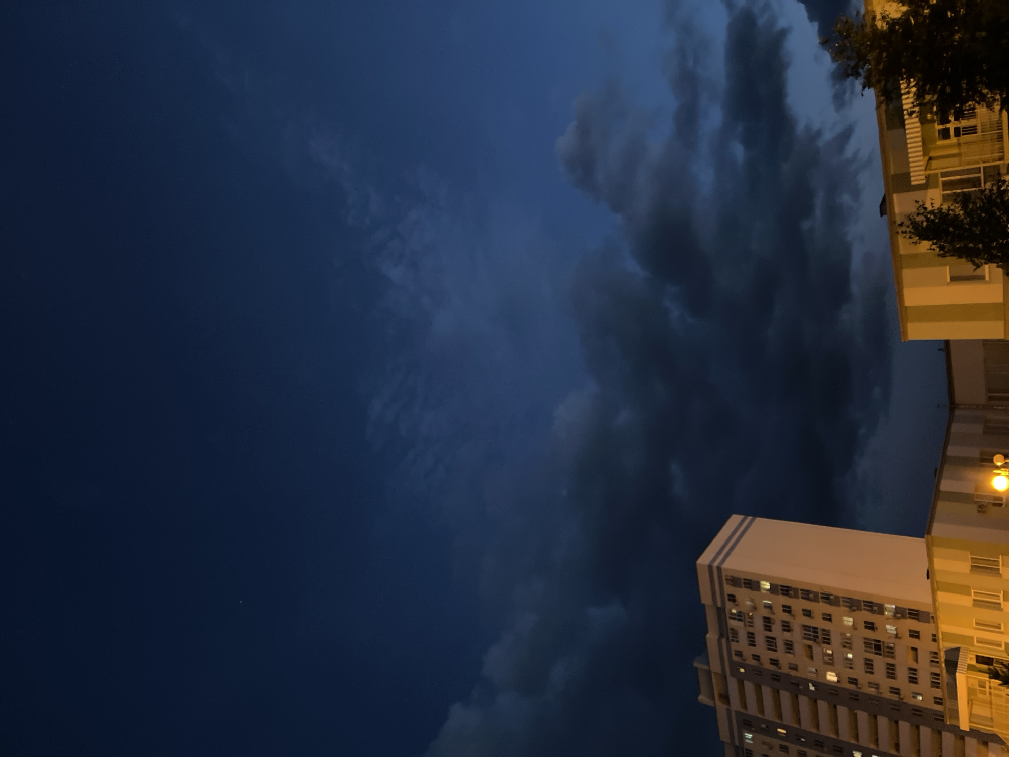 This media file depicts a nighttime scene in Novokuybyshevsk, Russia, dominated by a dark, cloudy sky. The image is oriented vertically, with the sky occupying the majority of the frame and a portion of a building visible on the right side.
The sky is a deep, dark blue, transitioning to almost black in some areas. Large, ominous storm clouds are present, with lighter patches suggesting some diffusion of light. A single, faint point of light, possibly a star or a distant light source, is visible in the lower left quadrant of the sky.
On the right side of the image, a multi-story building is partially visible. The building has a light-colored facade with horizontal bands of yellow and white. Numerous windows are visible, and many of them are lit from within, suggesting that people are present inside. The building appears to be an apartment complex or office building.
The time of day is clearly night, as indicated by the dark sky and the illuminated windows. The presence of storm clouds suggests potentially inclement weather, though no rain or lightning is explicitly depicted. The overall mood of the image is somewhat dramatic and possibly foreboding due to the dark clouds and the night setting. There is no visible text in the image.