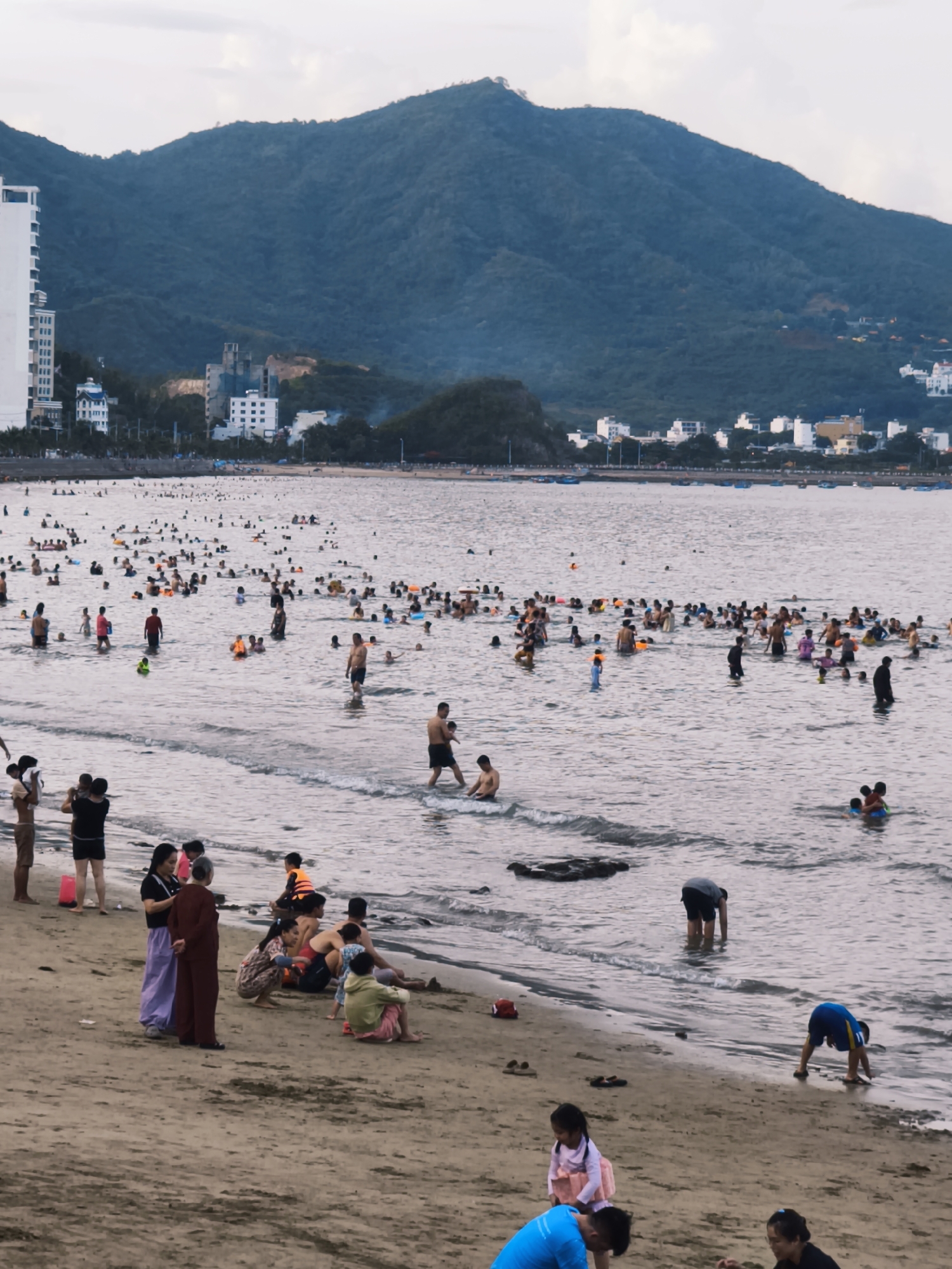 The image depicts a crowded beach scene in Nha Trang, Vietnam. A large number of people are in the water, swimming and playing. Some are wading near the shore, while others are further out. The water is calm with small waves. The beach itself is sandy, with some people sitting or standing on it. In the background, there are buildings and mountains. The sky is overcast. The overall impression is of a popular recreational area on a cloudy day.