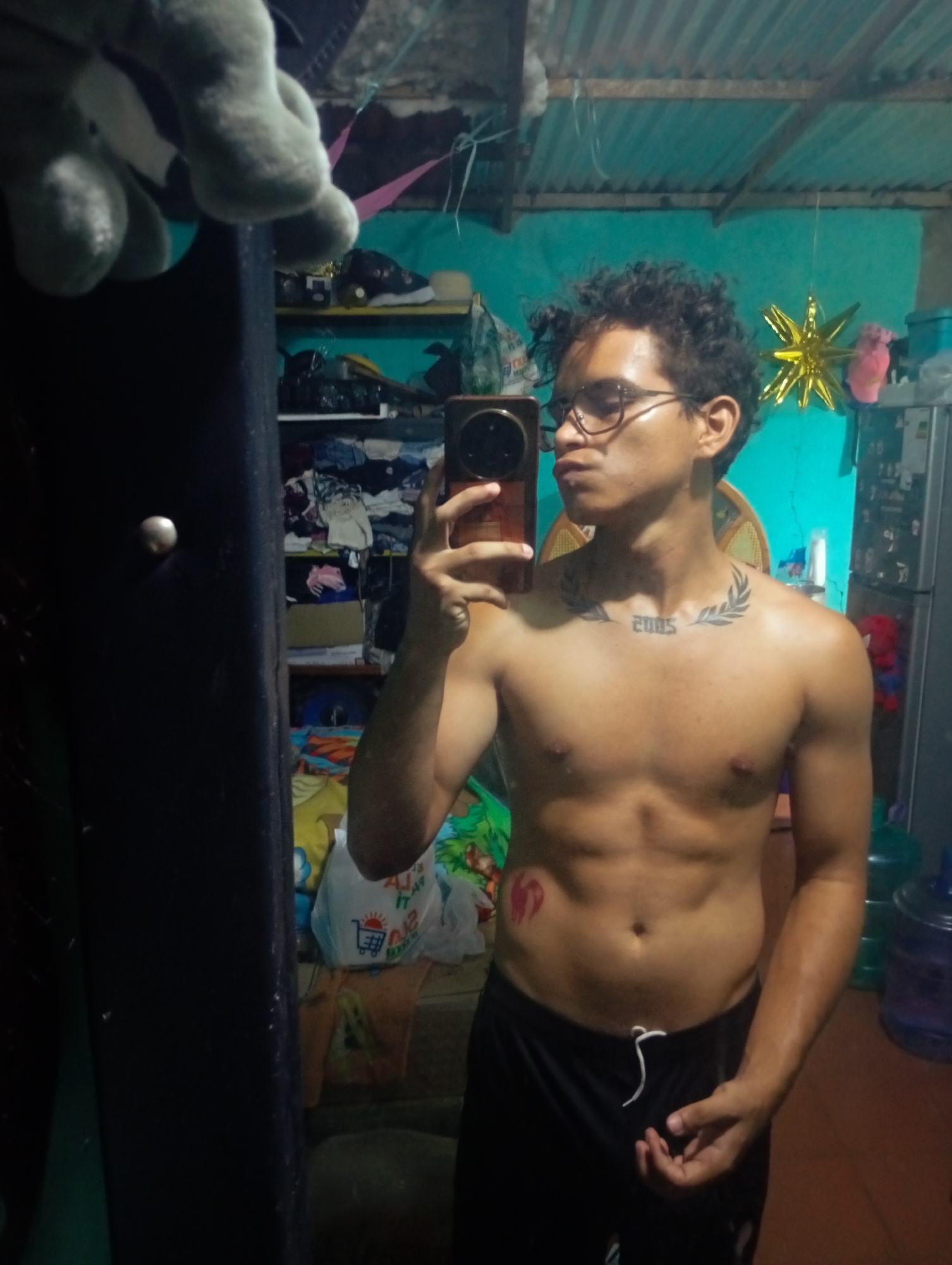 In Altagracia, Venezuela, a shirtless young man with curly dark hair and glasses is taking a mirror selfie in a cluttered room. He is wearing black athletic shorts with a white drawstring. He has a tattoo on his chest that appears to be laurel leaves with the numbers "2005" in the center, and a red mark, possibly a tattoo or drawing of a heart, on his left abdomen. The background reveals shelves filled with clothing and various items, a fan on the wall, and a large blue water jug. The lighting is dim, suggesting it is likely indoors, possibly a bedroom or living area in a residential setting. The presence of a plush toy in the foreground suggests a personal living space.