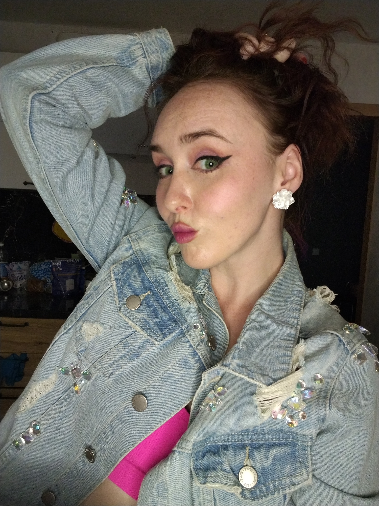 A young woman from Izhevsk, Russia, is captured in a close-up, indoor portrait. She poses playfully, holding her reddish-brown hair up in a messy bun or ponytail with her right hand, while pouting her lips towards the camera. Her green eyes are a prominent feature, enhanced by bold black winged eyeliner and pink eyeshadow. She wears bright pink lipstick and her fair skin shows visible freckles.
She is dressed in a light-wash denim jacket that is extensively distressed with rips and frayed edges, particularly around the collar, shoulders, and pockets. The jacket is embellished with numerous iridescent rhinestones and gems in shades of clear, pink, green, and blue, which sparkle under the lighting. A bright pink top or bra is visible underneath the open jacket. She accessorizes with a white, floral-shaped stud earring in her right ear.
The background is dimly lit and somewhat blurry, suggesting an indoor setting, possibly a home. Darker surfaces and indistinct shapes are visible on the left side of the frame. The lighting seems to come from the front, illuminating the woman's face and the sparkling details on her attire. Some of the jacket's metal buttons feature faint, unreadable text or a design. The overall impression is one of confidence and playful engagement.