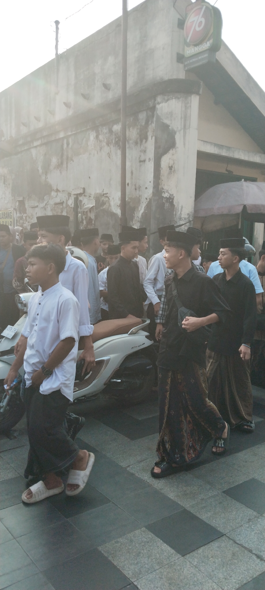 A group of young men are walking together in what appears to be an outdoor urban setting, possibly during a religious event. Most of the men are wearing traditional Indonesian clothing, including peci (caps) and sarongs. One person in the foreground is wearing a white collared shirt and black pants, while others are in black shirts and sarongs. A white scooter is parked to the left of the frame. The background shows a weathered, textured wall with some signs and an awning. The atmosphere is hazy, possibly due to sunlight or dust. The specific location is the city of Kauman, Indonesia, which is known for its rich history and Islamic heritage.
