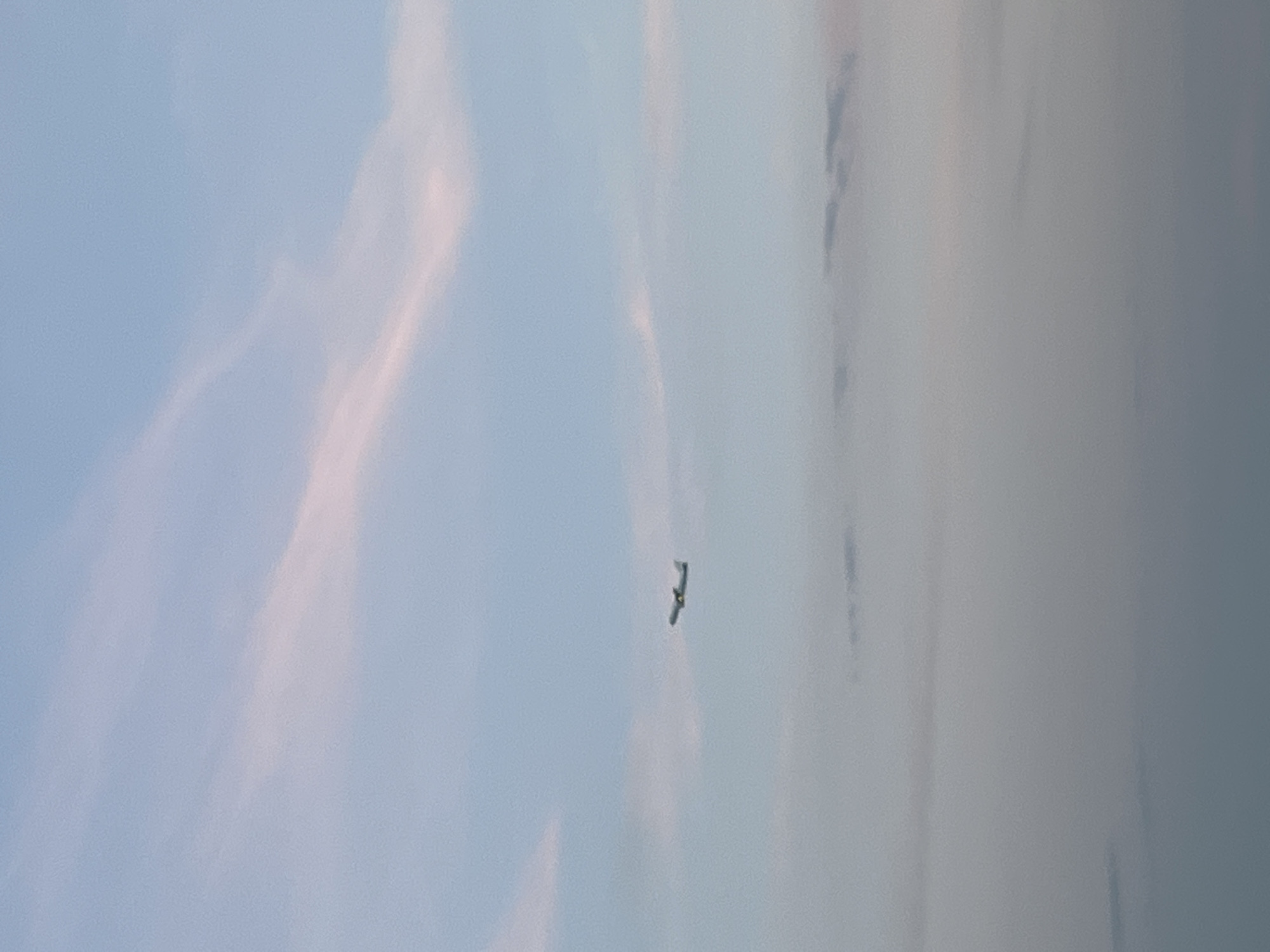 The image depicts a clear sky with some wispy pink and white clouds. In the middle ground, a small airplane is visible in flight, angled upwards as if climbing. The lower portion of the image is filled with a hazy, muted blue and grey layer, likely indicating clouds or mist at a lower altitude. There is no discernible indication of the city of Varna, Bulgaria, in this photograph.