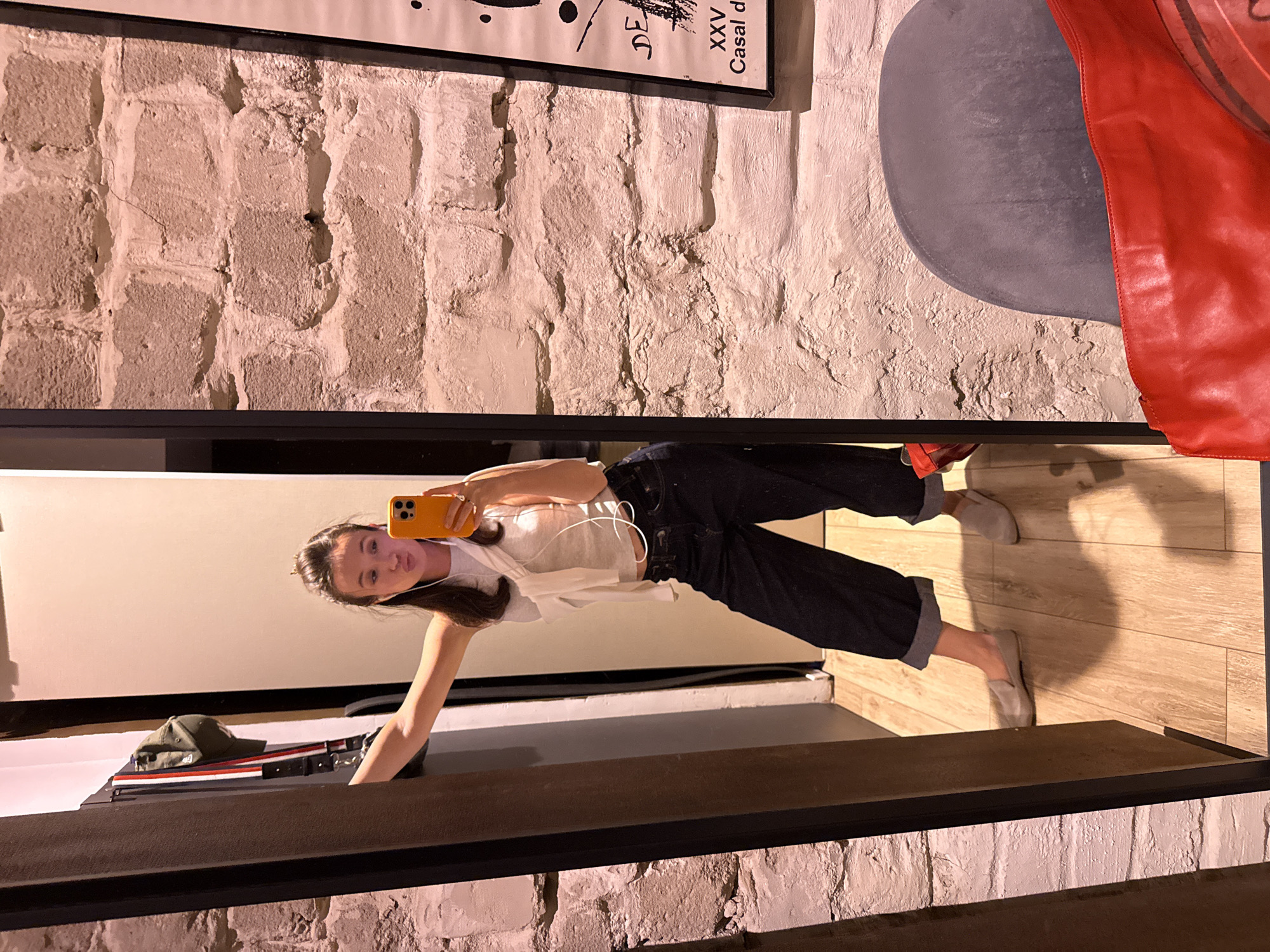 A young woman stands in what appears to be a fitting room, taking a selfie in a mirror. She is wearing a white crop top, dark wide-leg jeans, and light-colored slip-on shoes. She has earbuds in and is puckering her lips. To her left, a cap is hanging on a rack. The wall behind her is made of rough, textured bricks, and a poster is visible on the far right. The floor is made of wooden planks. The overall lighting is warm, suggesting artificial light. This scene is not specifically identifiable as being in Tel-Aviv, Israel, based on visual cues alone.