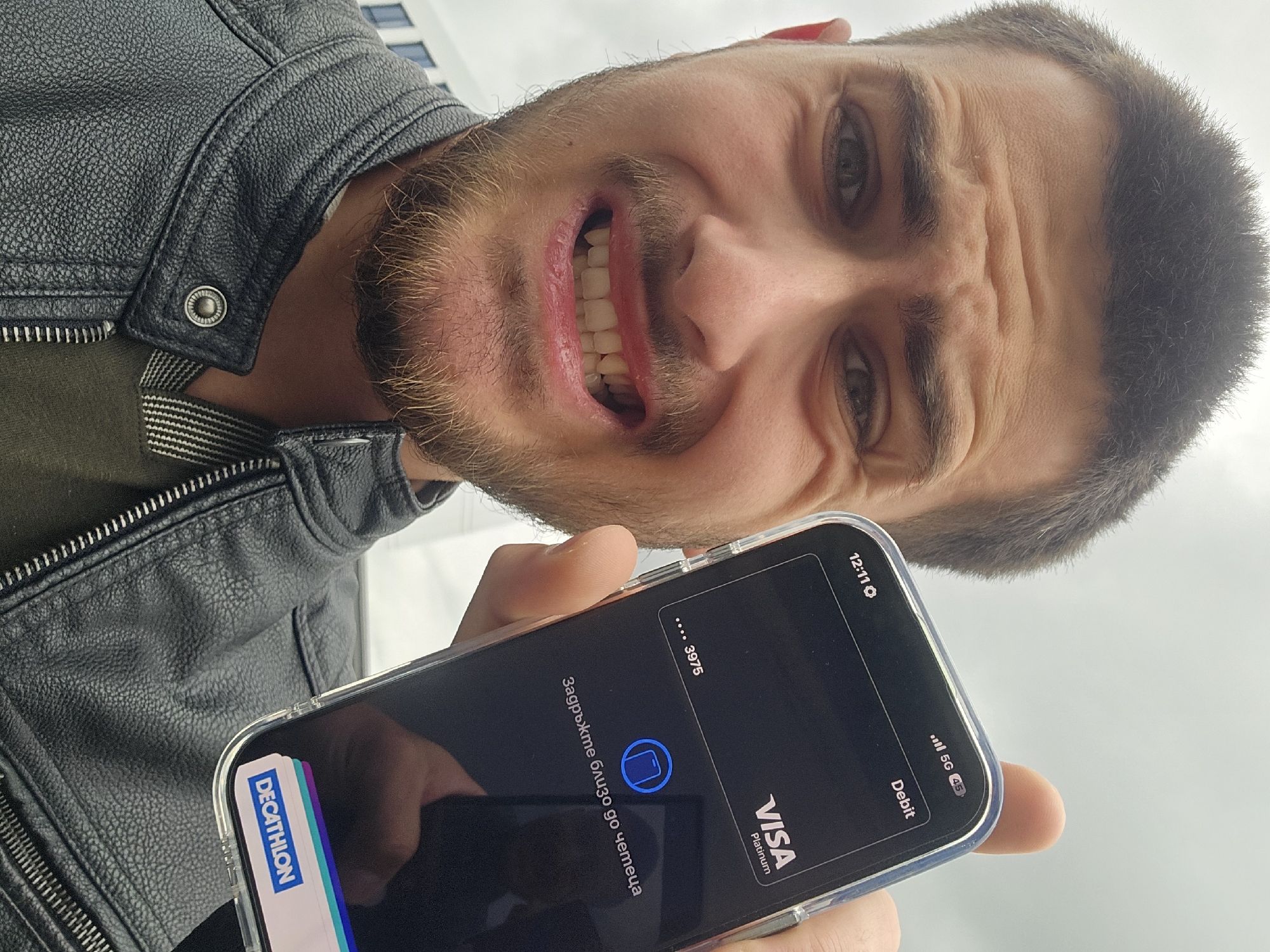 A man with a short beard and dark hair is captured in a close-up, low-angle shot, looking directly into the camera. He wears a black leather jacket with visible zipper and snap button details over a dark green shirt.
The man holds a smartphone in a clear case, displaying a digital payment interface, likely for a contactless transaction. A "DECATHLON" loyalty card or pass is visible tucked into the phone case. The phone screen shows a "VISA Platinum Debit" card with the last four digits "... 3975" and a prompt in Bulgarian: "Задръжте близо до четеца" (meaning "Hold close to the reader"), alongside a contactless payment icon. The time on the phone screen is 12:11, and a "5G" signal indicator is visible.
His facial expression is highly animated and exaggerated: his eyes are wide, his eyebrows are furrowed, creating deep wrinkles on his forehead, and his mouth is open in a wide, intense grimace, baring his upper teeth. This expression could convey frustration, surprise, or a playful, intense pose related to the payment process.
The background is a bright, diffuse grey, suggesting an overcast sky or a brightly lit indoor environment. The scene takes place in Essen, Germany, possibly at or near a Decathlon store given the visible branding.