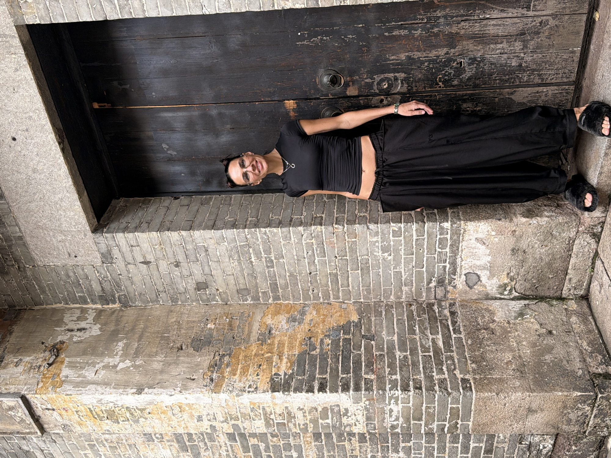 A woman stands in front of a dark wooden door in Chancheng District, China. She is wearing a black short-sleeved shirt that exposes her midriff, a necklace, and black pants with large pockets. She has on glasses and her hair is pulled up in two buns. The building is made of brick, with some sections appearing weathered and discolored. The woman is positioned to the left of the door, with her body angled slightly towards the viewer.