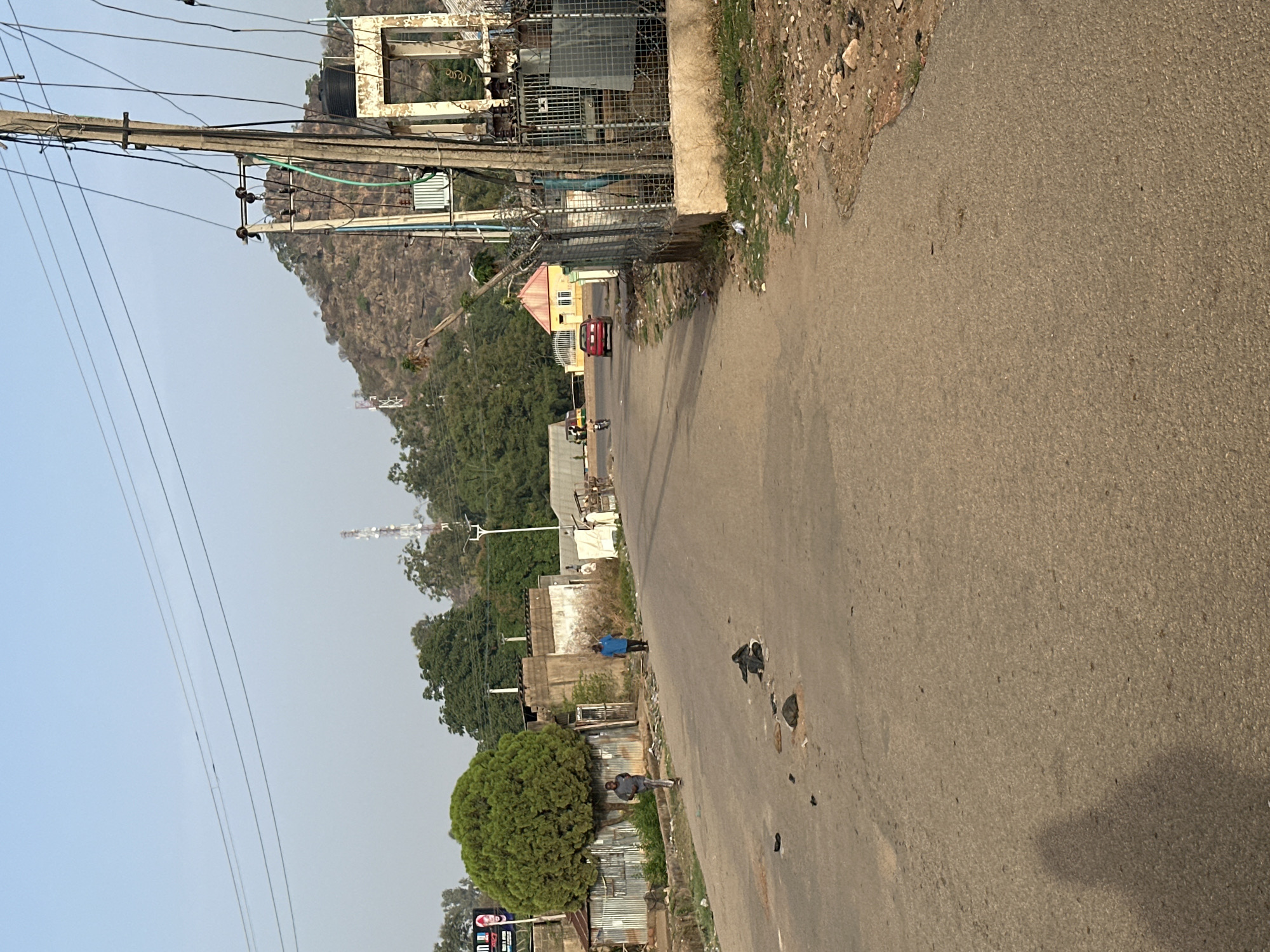 The image depicts a street in Bukuru, Nigeria. An asphalt road takes up most of the frame, with utility poles and wires running along the left side. Buildings, vegetation, and a hill are visible in the background. A red car is driving on the road in the background. People are standing or walking near the roadside. Garbage is scattered on the street.