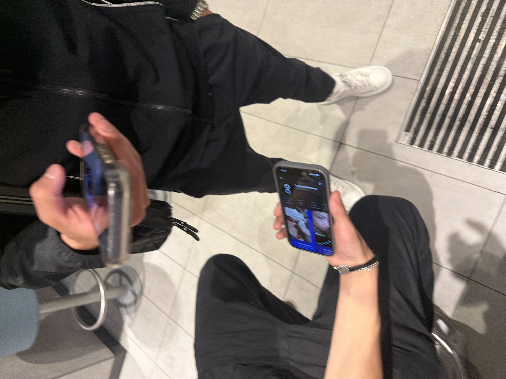 The image, captured from a high-angle perspective, depicts two individuals in an indoor setting, likely in Sofia, Bulgaria. Both are predominantly dressed in black attire, consisting of black pants and tops/jackets, and are wearing white sneakers. The flooring is light-colored tile, and a dark striped mat or grate is visible on the right side of the frame.
The individuals are focused on their smartphones. The person on the right holds their phone vertically, displaying an application that appears to be a video call or a social media feed with multiple faces. The word "Go" is visible at the bottom of this phone's screen, and the number "8" is seen in the top left corner, possibly indicating a timestamp or battery level. This individual also wears a metallic watch on their left wrist. The other person, mostly visible from the waist up, also holds a smartphone, though its screen is largely obscured by their hand. A black bag strap is seen near this person's left hip.
A metallic footrest or part of a chair/stool is partially visible near the feet of the person on the left, suggesting they might be seated. The overall impression is one of people occupied with their devices in a public or semi-public indoor space.