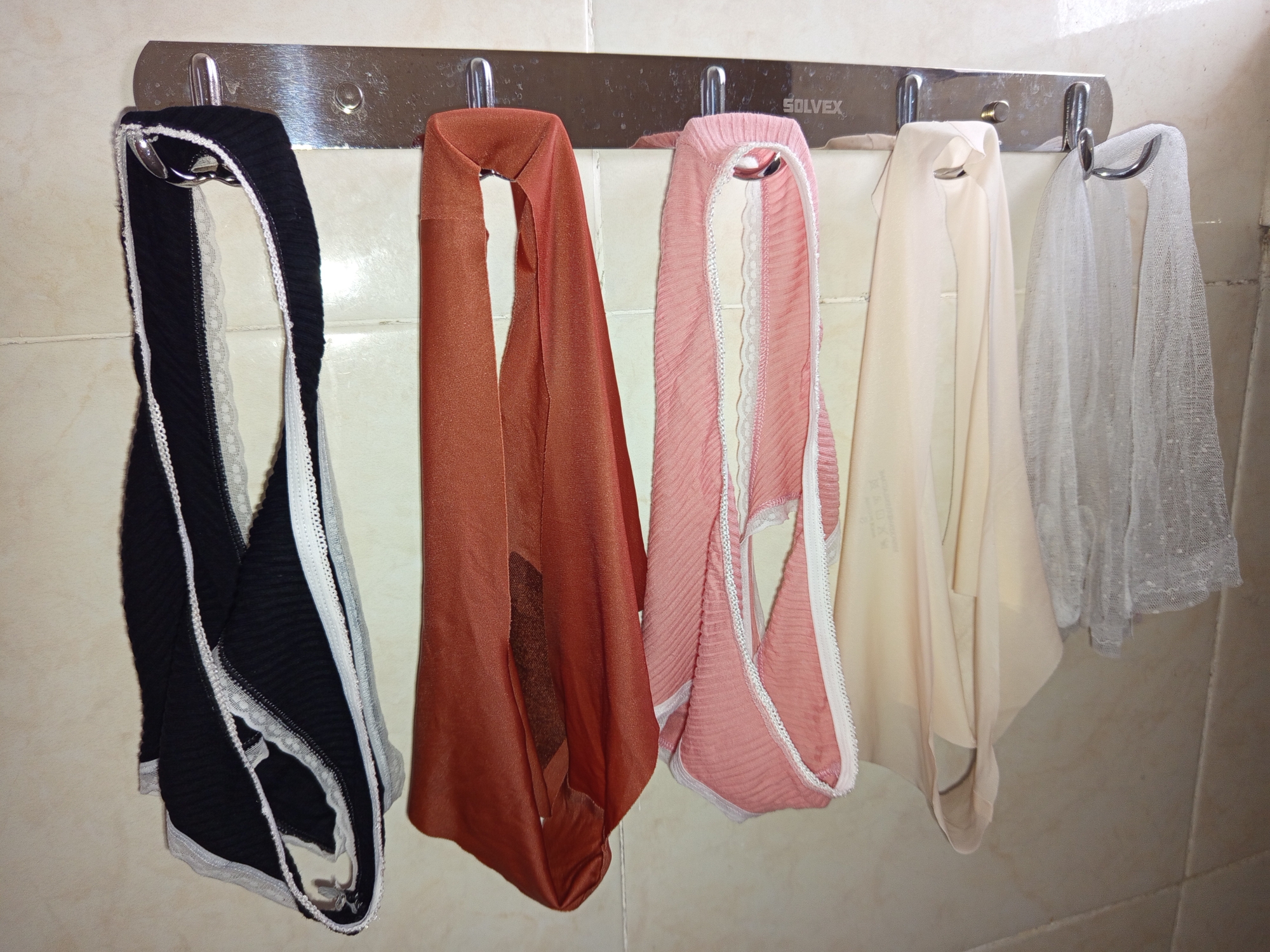 A stainless steel wall-mounted rack with five hooks displays women's underwear. The rack is mounted on a tiled wall. From left to right, the underwear are black with white trim, rust-colored, pink with white trim, light beige, and white lace. The word "SOLVEX" is visible on the rack. The location is North Kuta, Indonesia.