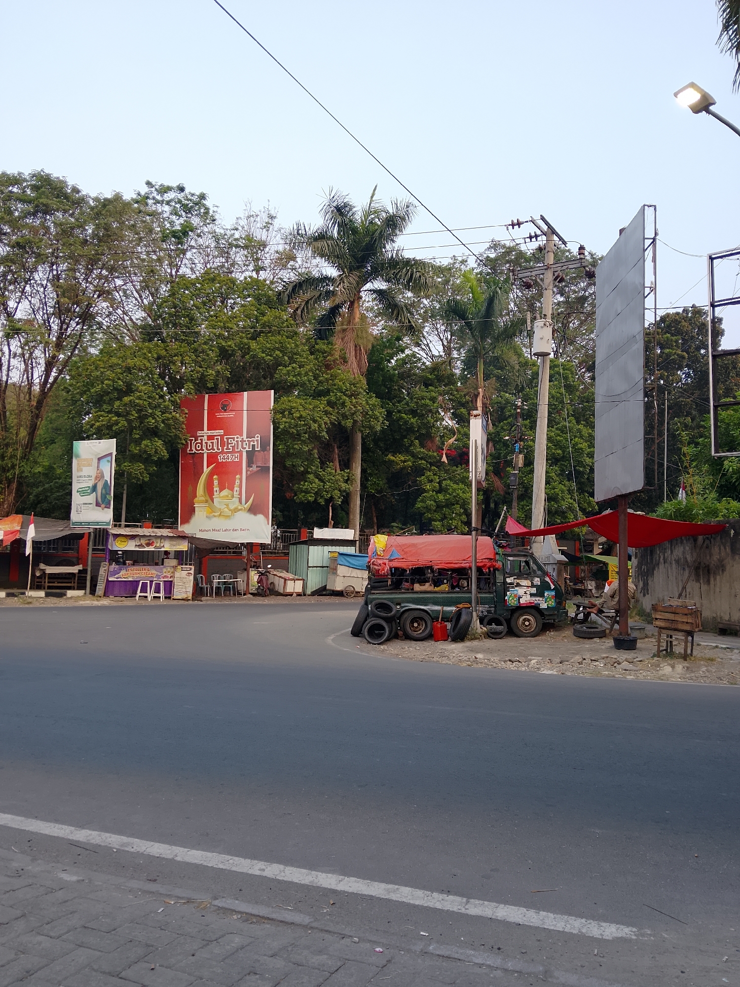 This image depicts a roadside scene in Balun, Indonesia, featuring a small collection of shops and a parked truck. In the foreground, a paved road with a white lane marker runs horizontally. To the left of the road, a sidewalk is constructed with interlocking gray pavers.
Dominating the background is a dense arrangement of lush green trees, including palm trees, creating a natural backdrop. A clear, pale blue sky is visible above the trees. A power pole with insulators and wires stands prominently on the right side of the image, alongside a tall, grey billboard. A street lamp is illuminated and positioned on an arm extending from the power pole.
In the middle ground, several small businesses are situated along the roadside. One building, painted red and white, displays a large banner with "Idul Fitri 1447H" and an illustration of a mosque and crescent moon. Next to it, a smaller green banner features a woman and text. Further to the left, another shop appears to be selling food, with tables and chairs set up outside.
A green truck with a red tarp covering its cargo is parked on the right side of the scene, facing left. Several tires are stacked on the ground near the truck, suggesting it might be a tire repair or sales establishment. A red fuel can is also visible next to the tires. A small wooden crate sits on the ground near the truck. A person, partially obscured, is seated on a stool to the right of the truck. Red awnings are extended over some of the shops and the truck, providing shade. A small Indonesian flag is attached to a pole behind one of the shops. The overall impression is a vibrant, bustling commercial area situated along a main road surrounded by abundant greenery.