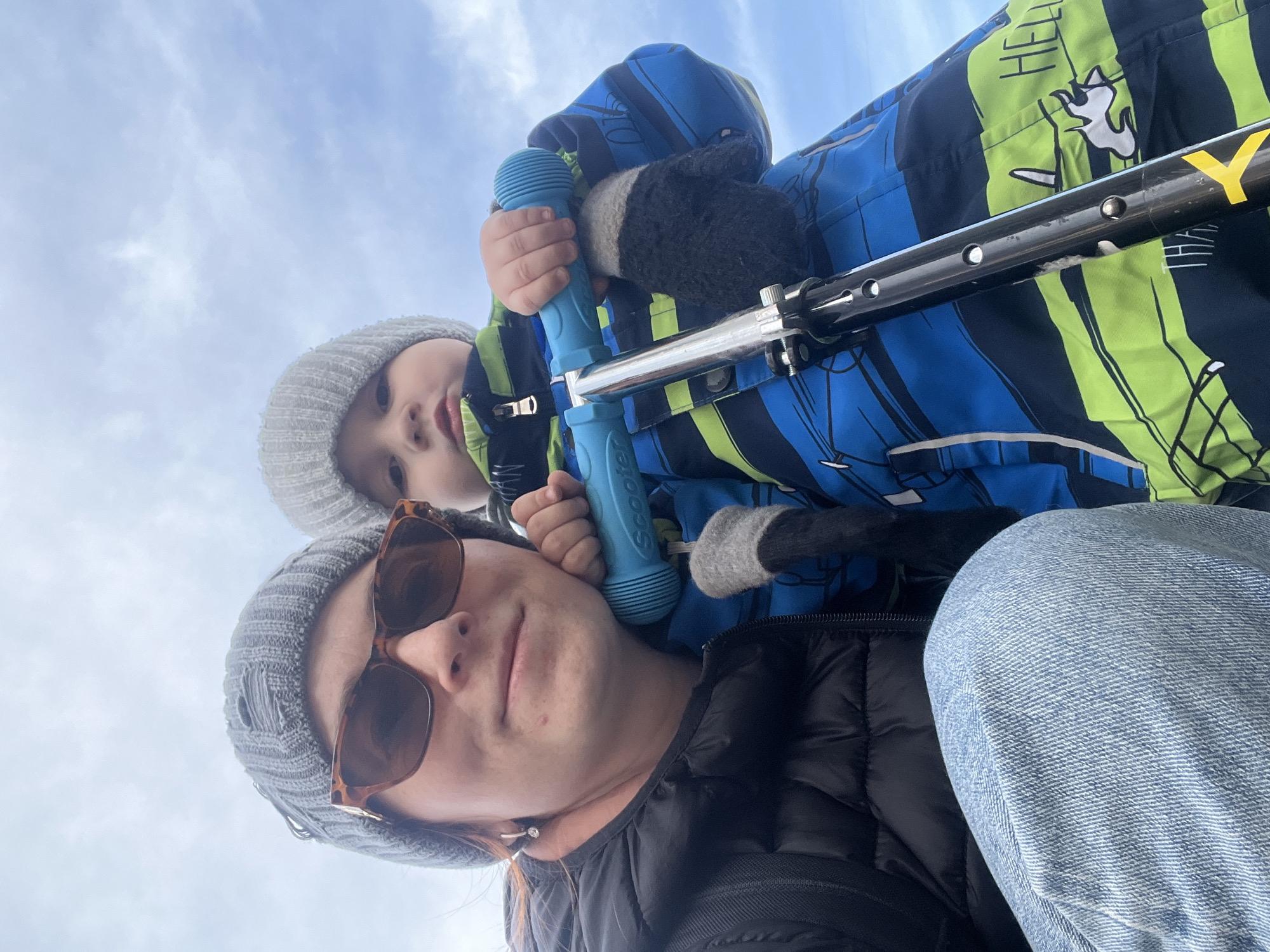 Here is a detailed description of the image:
**Scene**: A selfie-style photograph looking upwards, showing a woman and a child outdoors. The background shows a cloudy sky.
**People**: A woman wearing a dark puffer jacket, a gray beanie, and sunglasses is at the bottom of the frame. A child wearing a patterned winter jacket and a gray beanie is situated higher in the frame.
**Objects**: The child is holding a blue toy microphone. A black and silver scooter is positioned between the woman and the child.
**Actions/Interactions**: The woman is holding the child, who is holding a toy microphone, and the scooter. They are seemingly posing for a selfie.
**Environment**: The setting appears to be outdoors, likely during the day, as indicated by the presence of sunlight and a cloudy sky.
**Location**: While the image itself does not contain explicitly locational features to determine where it was taken, there is nothing to indicate the city of Chaikovsky, Russia.