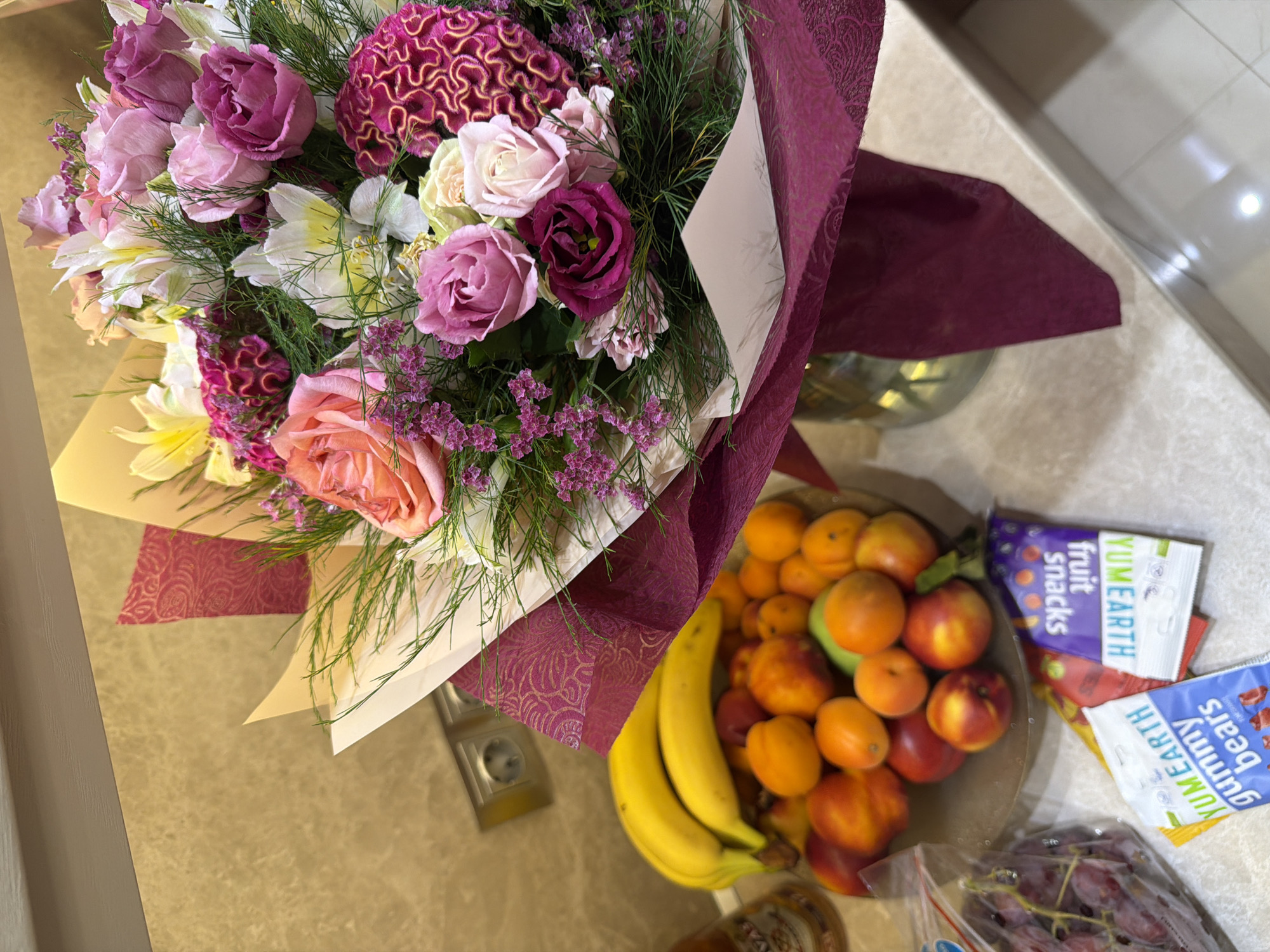 A bouquet of assorted flowers, including roses and lilies in shades of pink, purple, and white, is displayed on a counter. Next to the bouquet, a bowl of mixed fruits, including bananas, peaches, and tangerines, is visible. Also present are a bunch of red grapes, and two small packages of "YUMEARTH" gummy bears and fruit snacks. The background shows a beige-tiled wall with electrical outlets. The scene is likely indoors, possibly in a kitchen or living area. There are no people or animals in the image. The lighting appears to be artificial. The overall impression is one of a domestic setting, with a focus on a gift of flowers and a selection of fruits and snacks. The location in Sofia, Bulgaria, is not evident from the visual information.