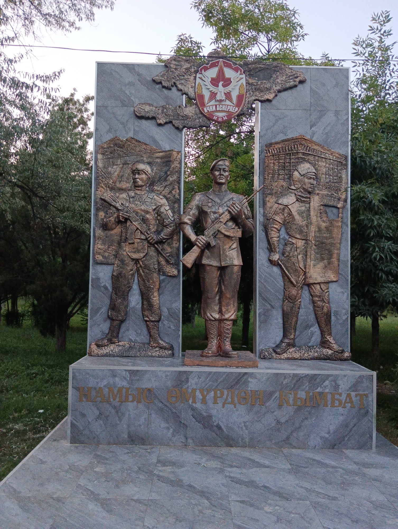 An outdoor memorial monument is depicted, featuring three bronze figures of soldiers in relief against a grey stone backdrop. The figures are positioned in a triptych format, with flanking figures of uniformed soldiers holding rifles, and a central figure of a soldier also holding a rifle. Above the central figure, a crest with a red star and a bird is visible. Below the figures, an inscription in gold lettering reads "НАМЫС ӨМҮРДӨН КЫМБАТ" which translates to "Honor is more valuable than life". The monument is situated in a grassy area with trees in the background. The sky is overcast.