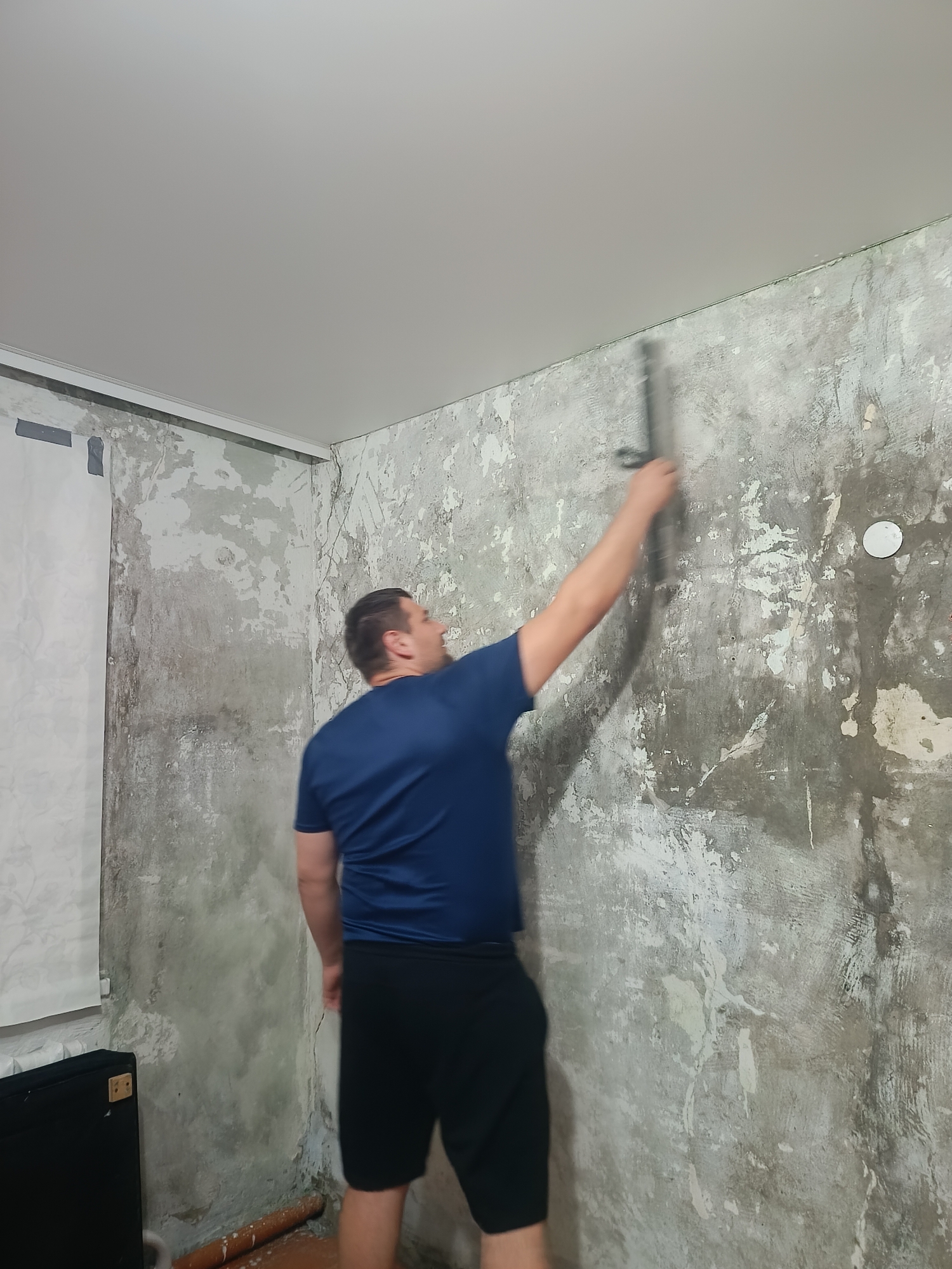 A man is standing in a room and scraping a wall with a tool. The room is under renovation. The walls are bare concrete, and the ceiling is white. The man is wearing a blue t-shirt and black shorts. The room is dimly lit.